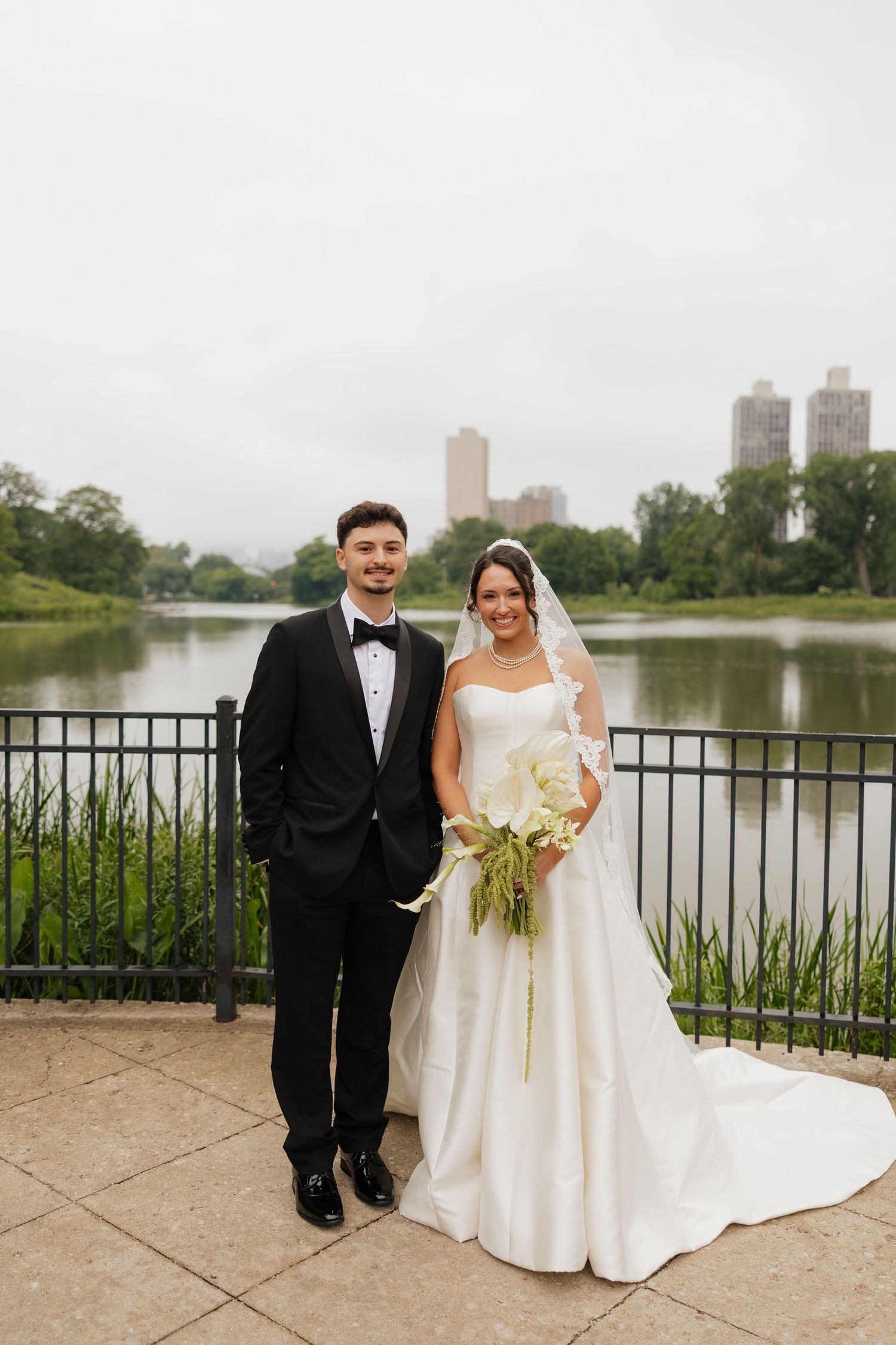 Bride and groom posing by a lake in Central Park, NYC, bride holding white bouquet in elegant gown and veil.