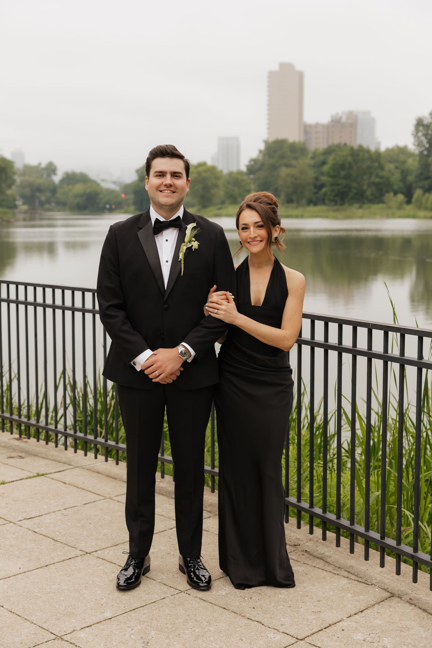 Couple in matching black formal attire posing by a lakeside railing with city skyline in background.
