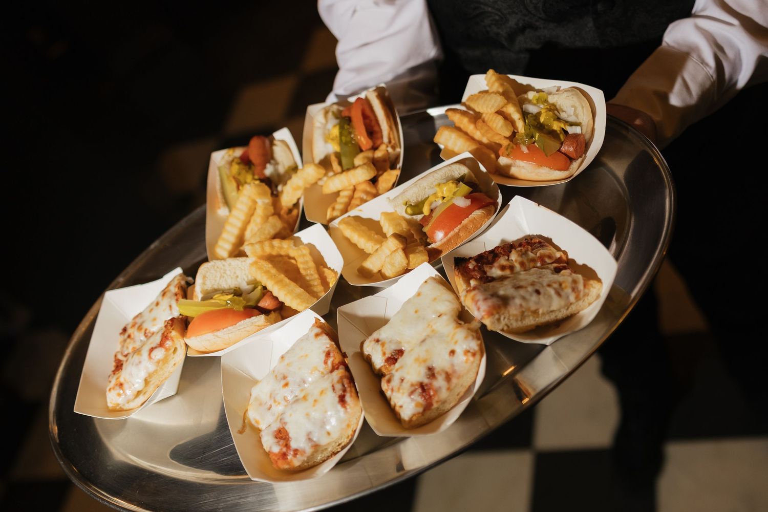 A server holds a tray of assorted sandwiches and french fries served in paper holders at a catered event.