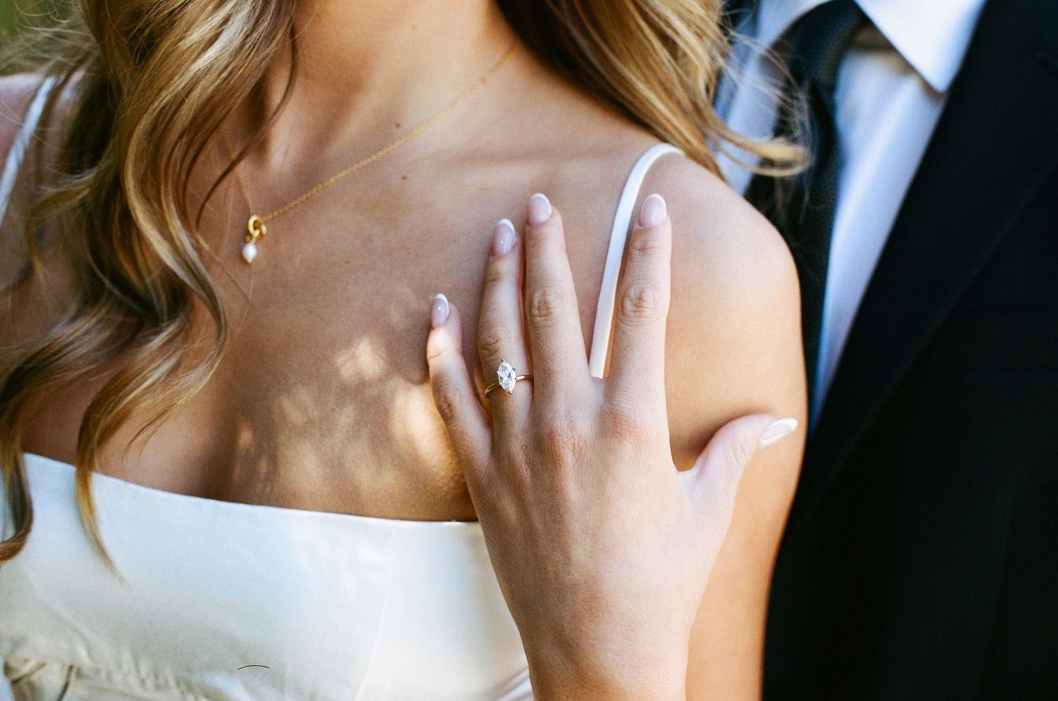 Bride showcasing elegant diamond engagement ring on hand resting on chest, wearing white gown and gold necklace.