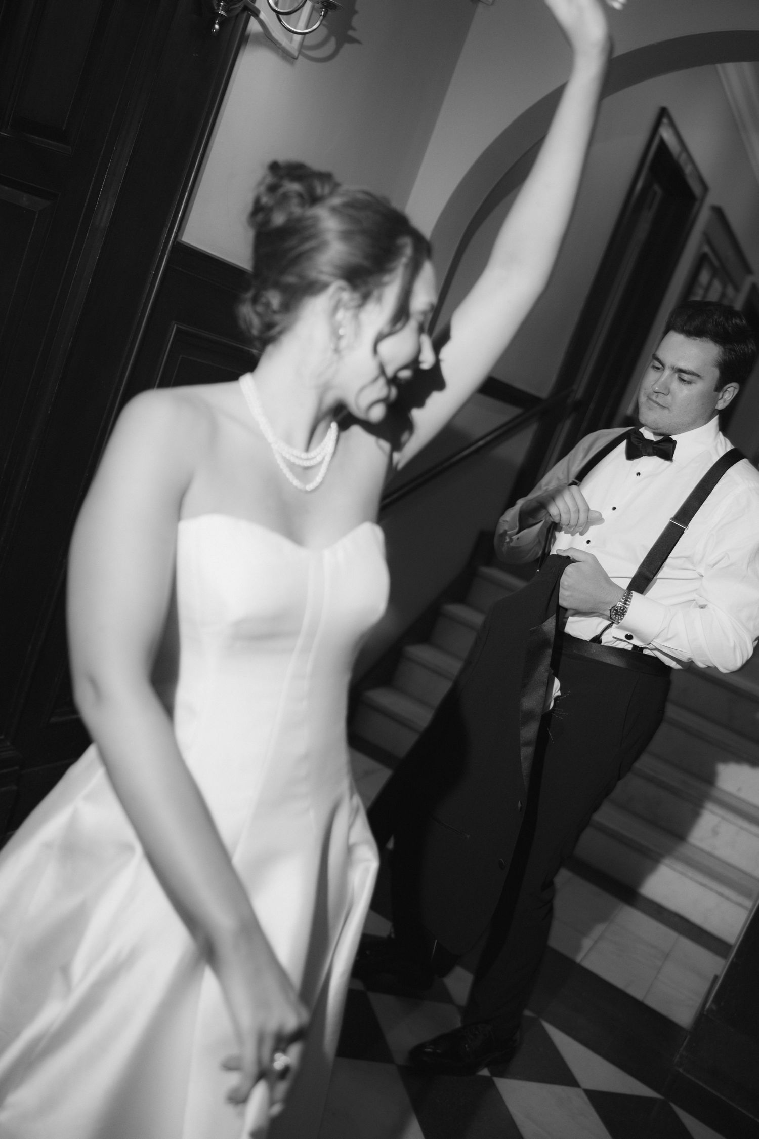 Black and white photo of a bride in strapless gown dancing while groom in suspenders watches during wedding reception.