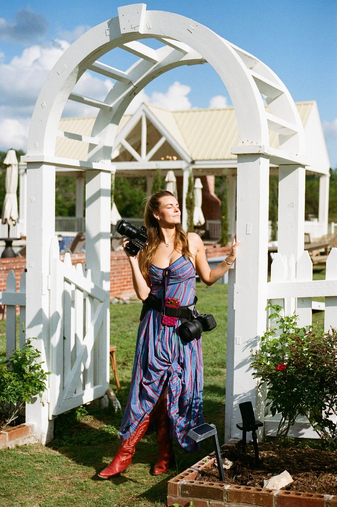 Woman in blue dress and red boots holds camera under white garden arch on sunny day.
