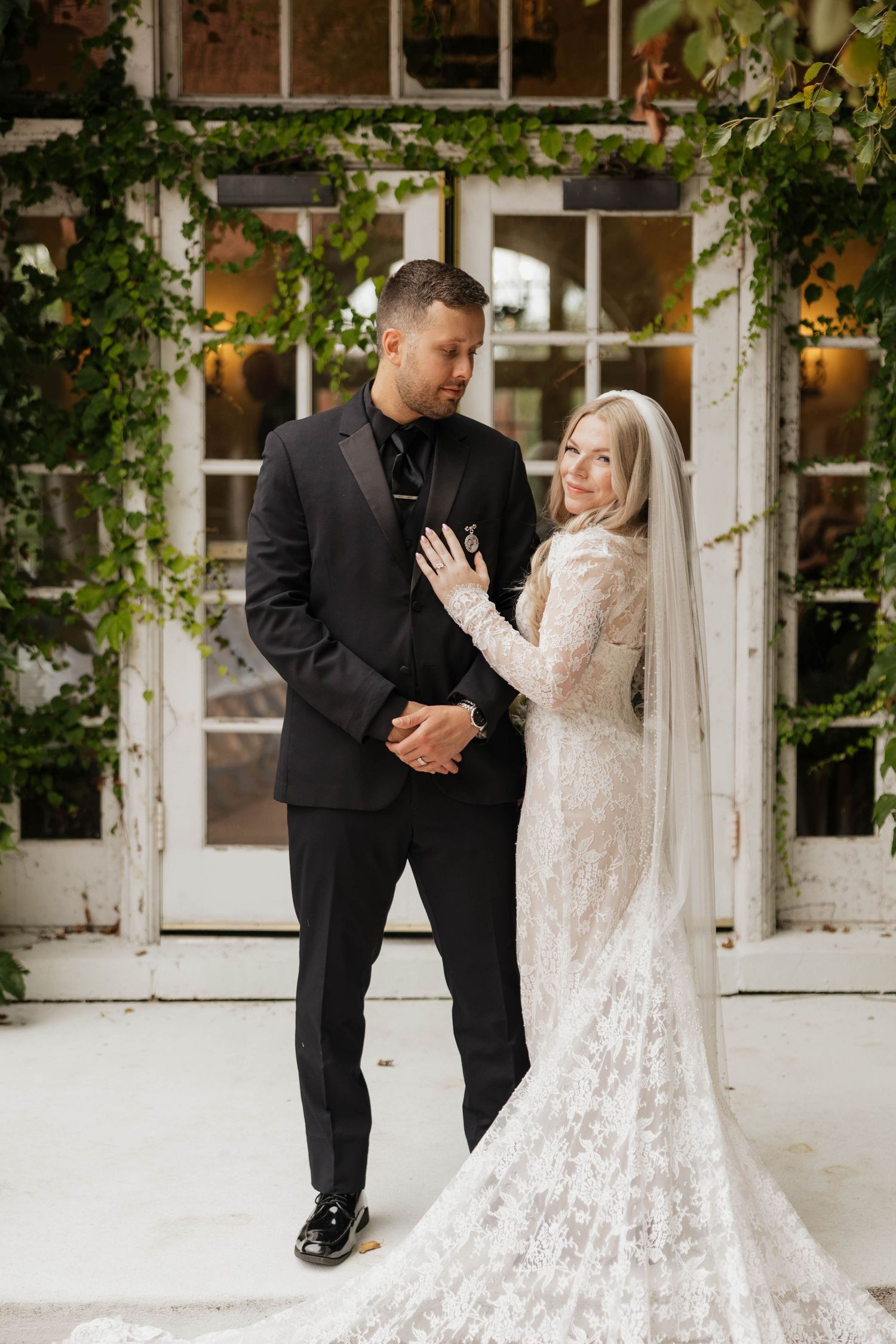 Bride in a long-sleeved lace wedding gown and veil poses with a groom in a black suit in front of white French doors covered in ivy.