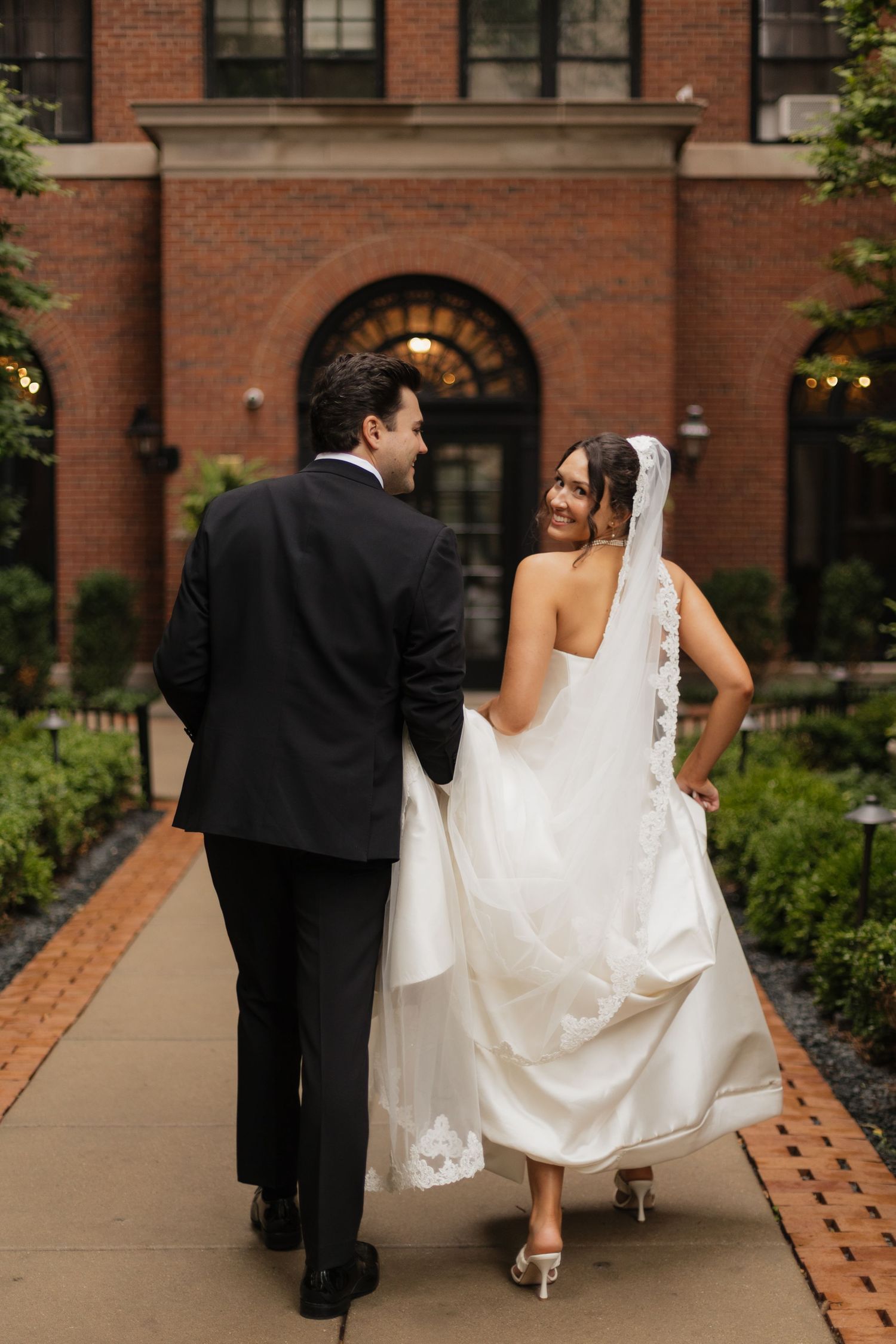 Bride in a white gown with lace-trimmed veil and groom in a black suit walk arm in arm toward a red brick building.
