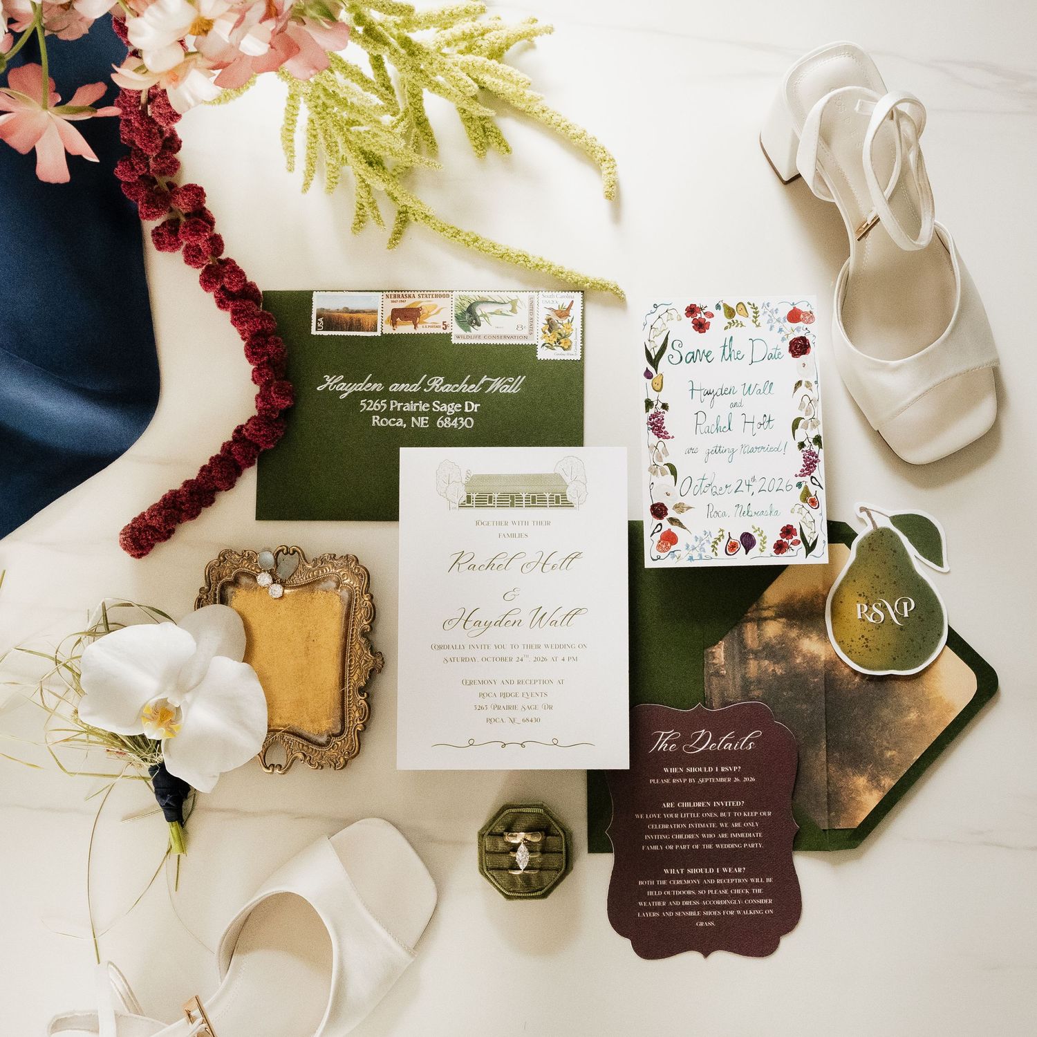 Flat lay of a green and floral wedding invitation suite with envelope, details card, pear-shaped tag, white heels, orchid, and ring box.