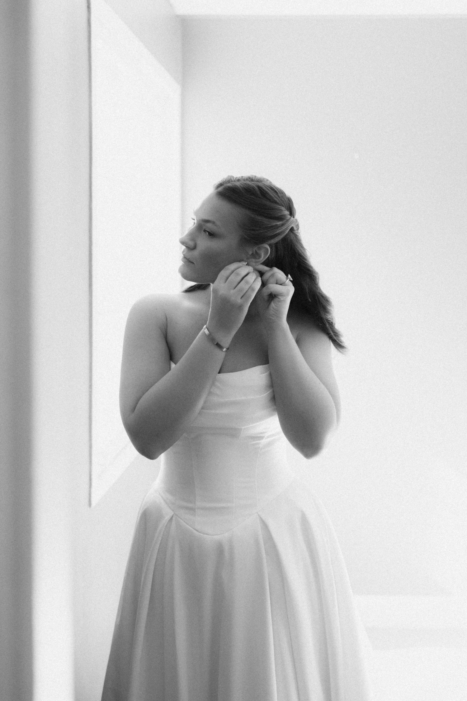 Black and white portrait of a bride in a strapless gown adjusting her earring beside a softly lit window.