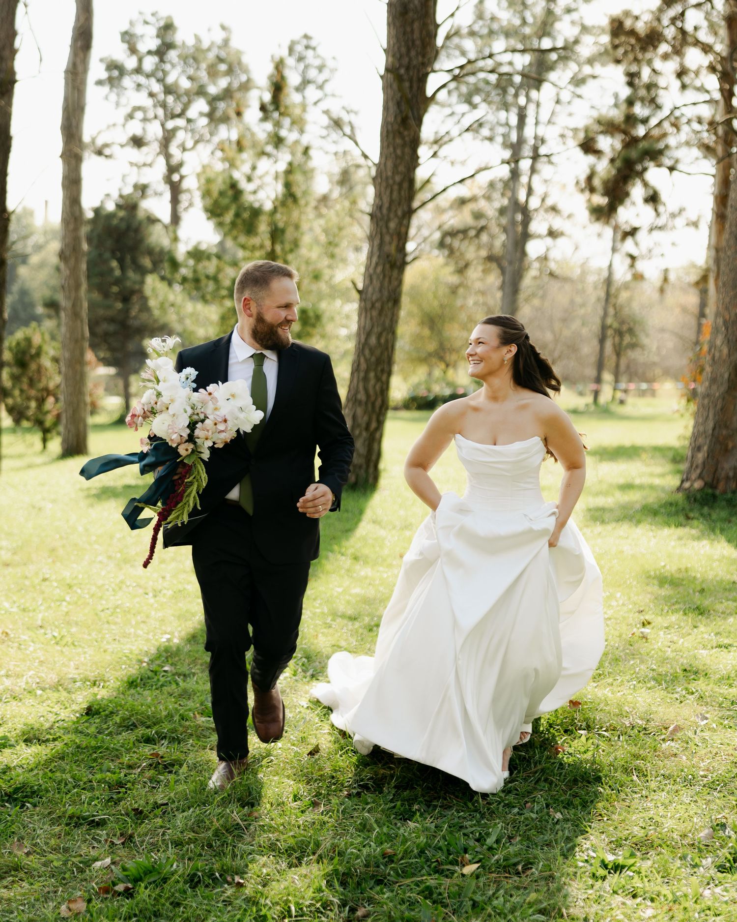 A smiling bride in a strapless white gown and a groom in a dark suit holding a bouquet walk hand in hand across a sunny grassy park.