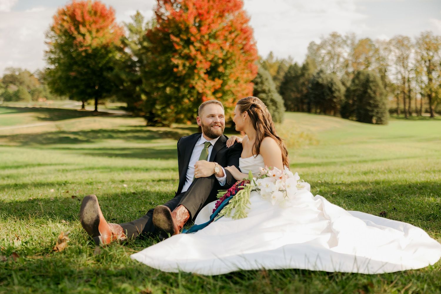 Bride in a white gown and groom in a dark suit sit close together on a grassy field with colorful autumn trees in the background.
