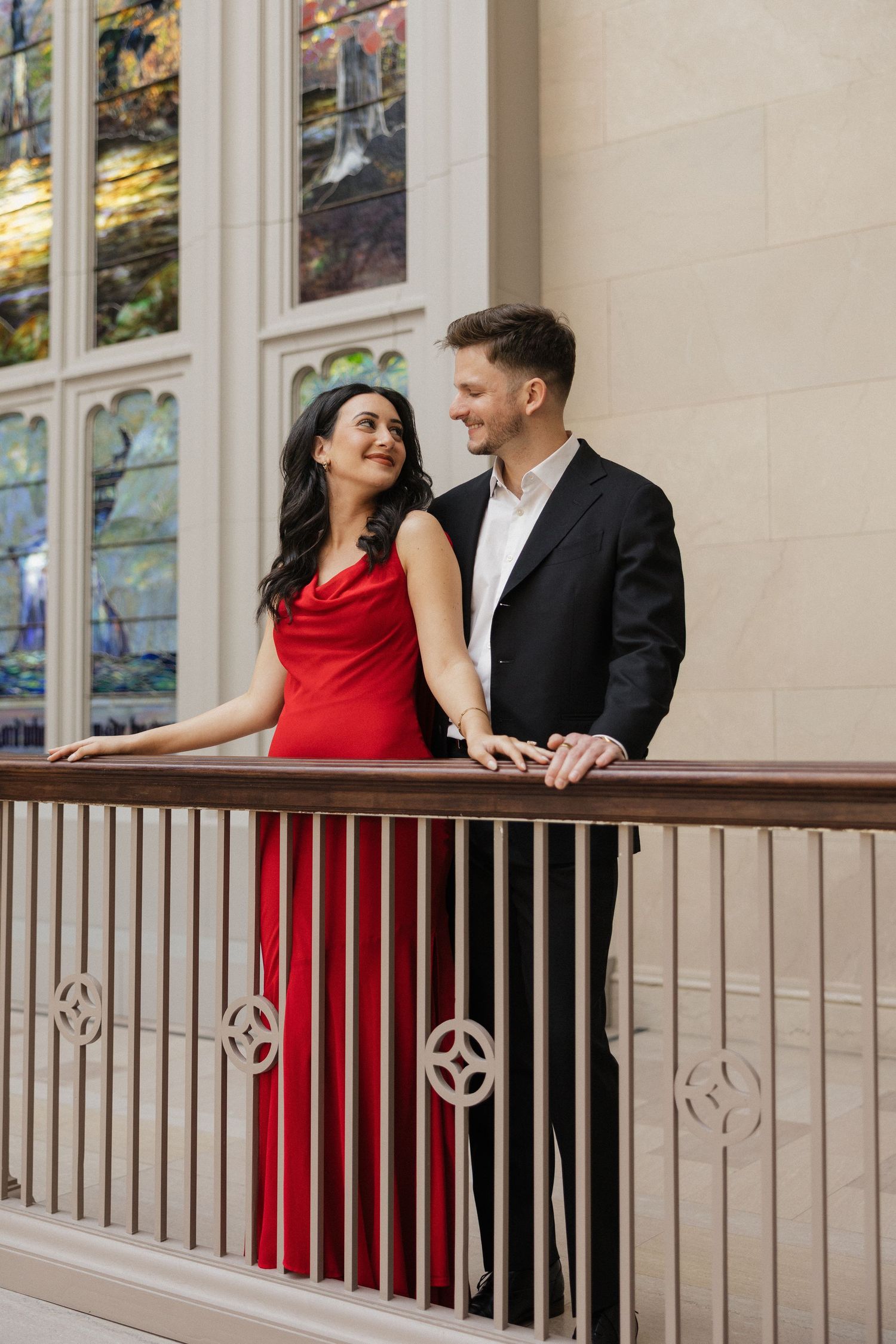 A couple shares a romantic moment on a balcony with stained glass windows, the woman in red and man in black suit.
