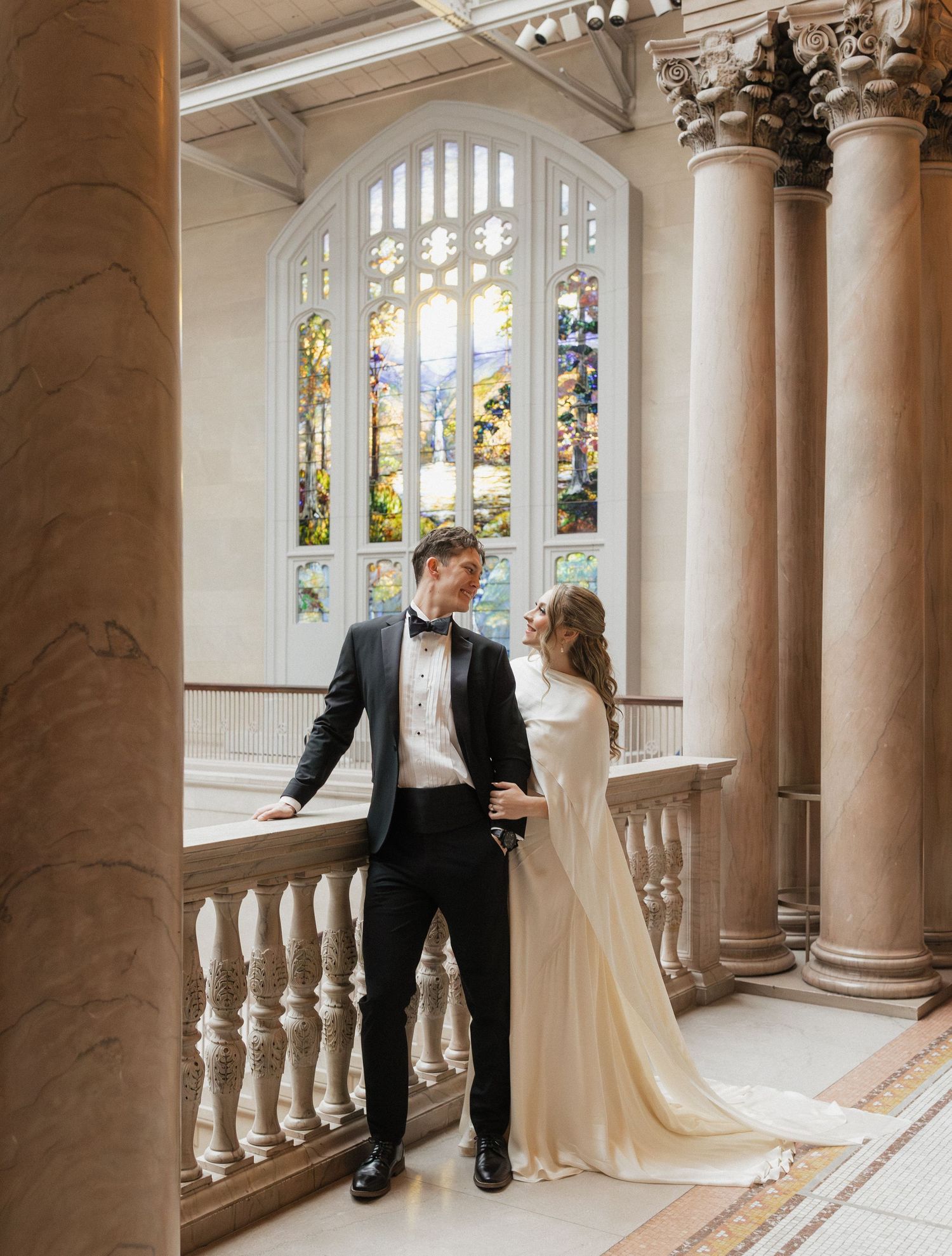 A couple shares a romantic moment in an elegant hallway with ornate columns and stained glass windows.