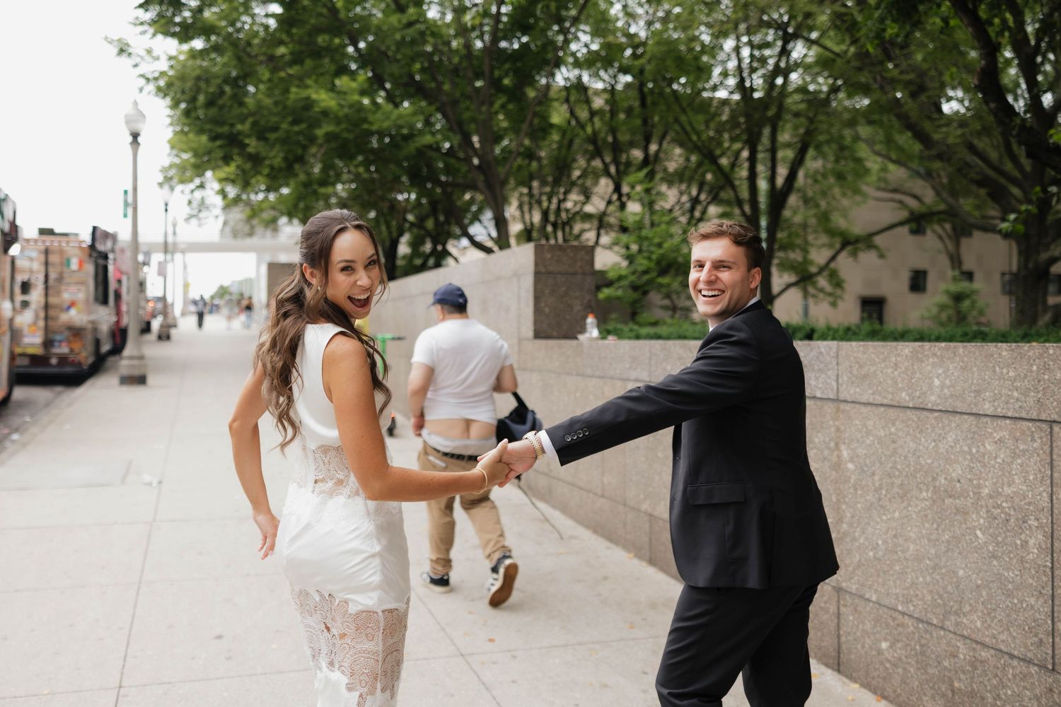 Couple in formal attire holding hands on a city sidewalk, woman in white dress looking back, man in black suit smiling.