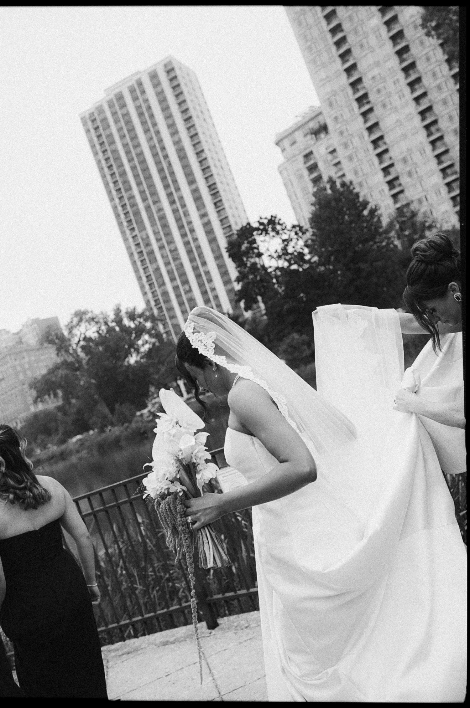 Bride holding bouquet with bridesmaids adjusting veil near city skyline.