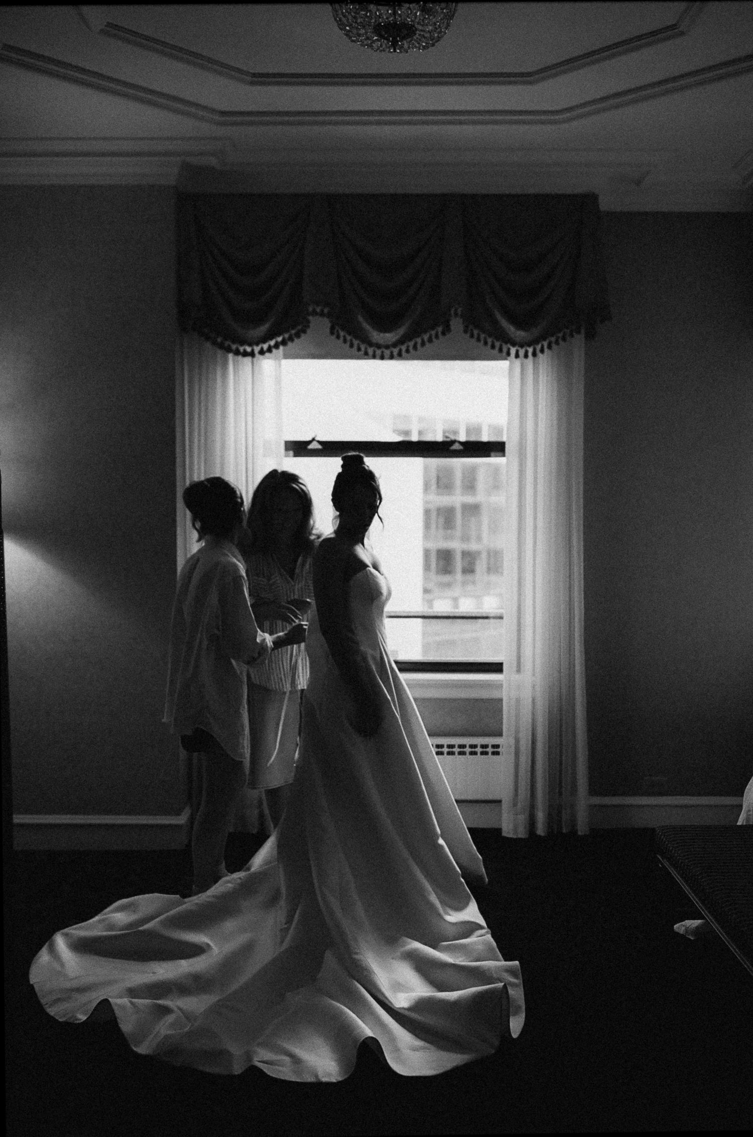 Silhouette of a bride in a flowing wedding gown being helped by attendants near a draped window.