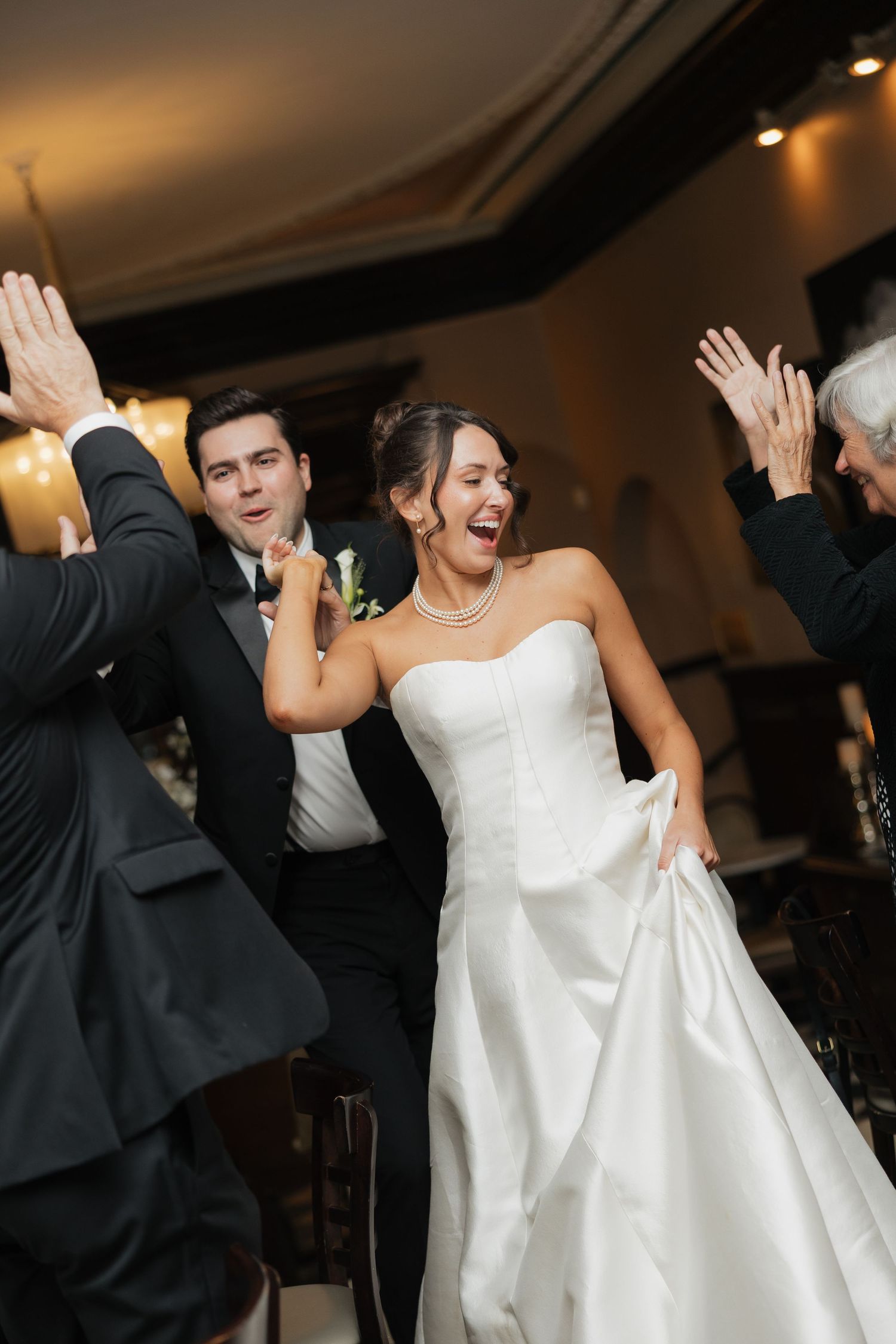 Joyful bride in white strapless gown and groom in black tuxedo making grand reception entrance with cheering guests.