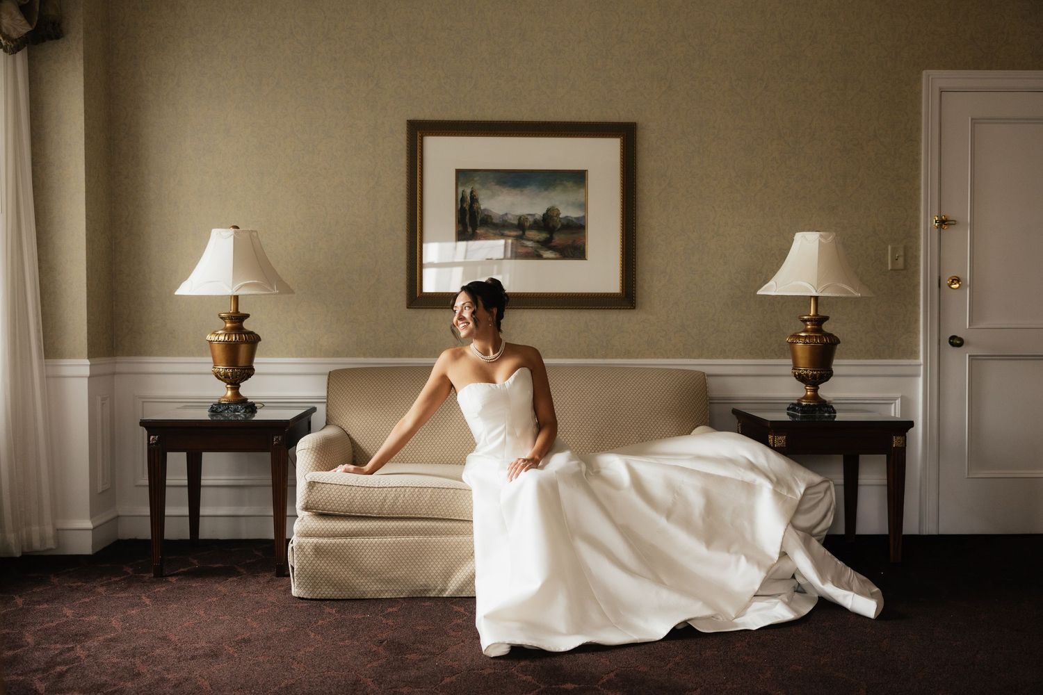 Elegant bride in a white strapless gown sitting gracefully on a cream sofa in a warmly lit hotel suite.