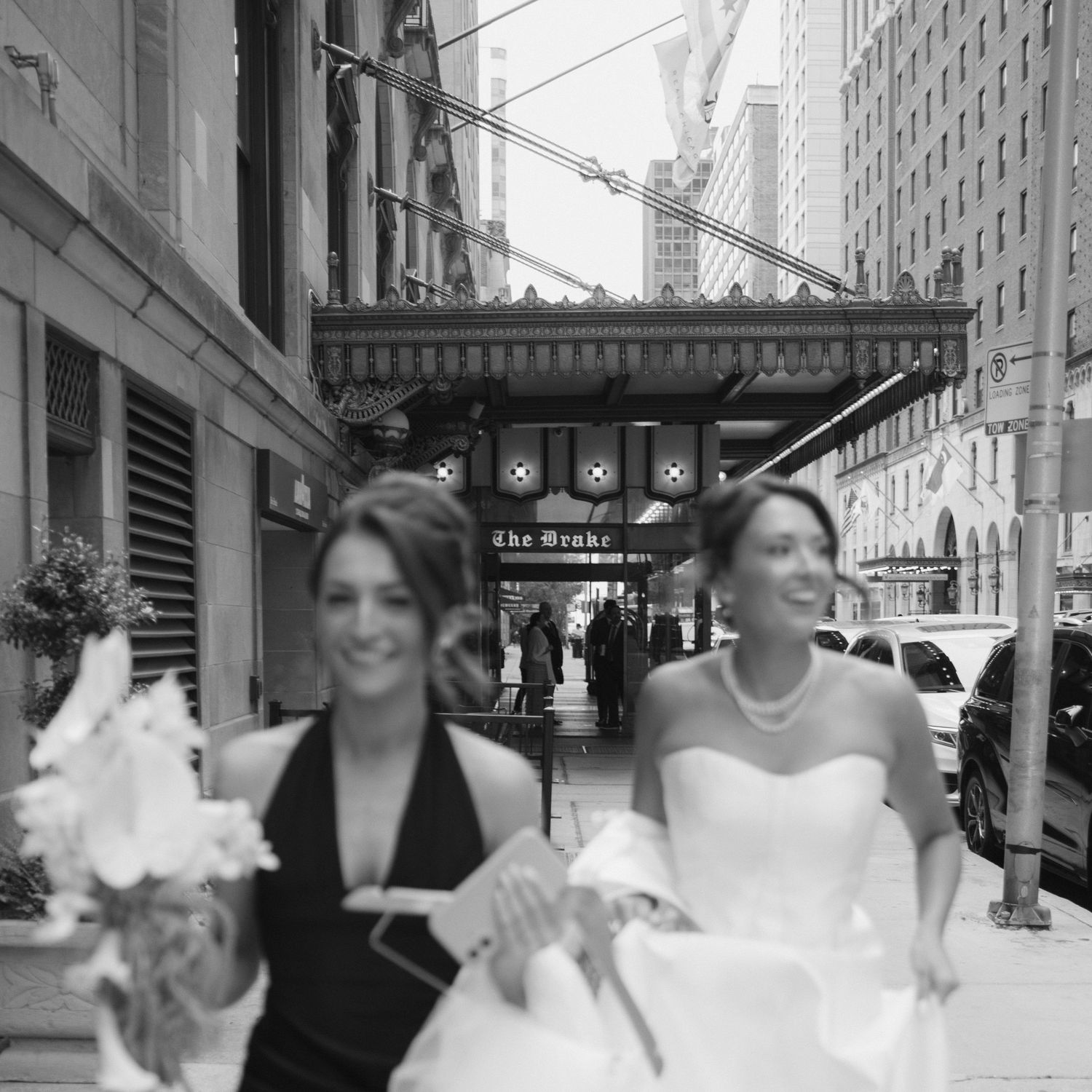 Black and white photo of two women in formal attire walking on a city street, one in a wedding dress holding flowers.
