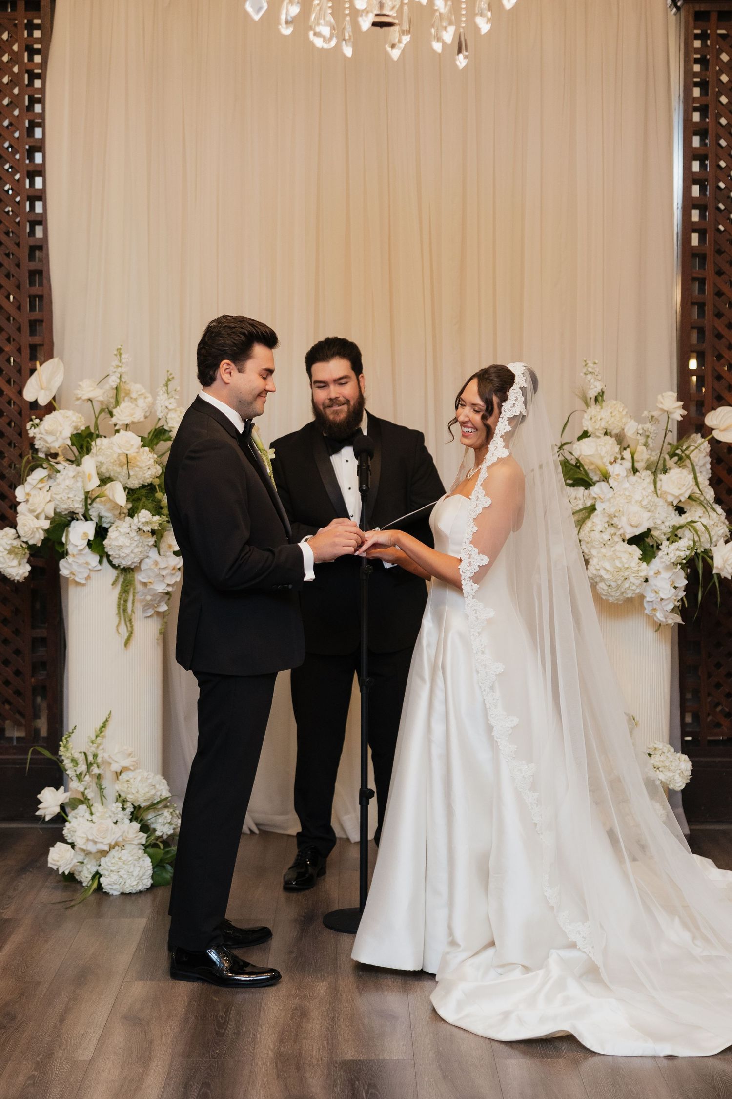 Groom places wedding ring on bride's finger during elegant indoor ceremony with white floral arrangements and cream drape backdrop.