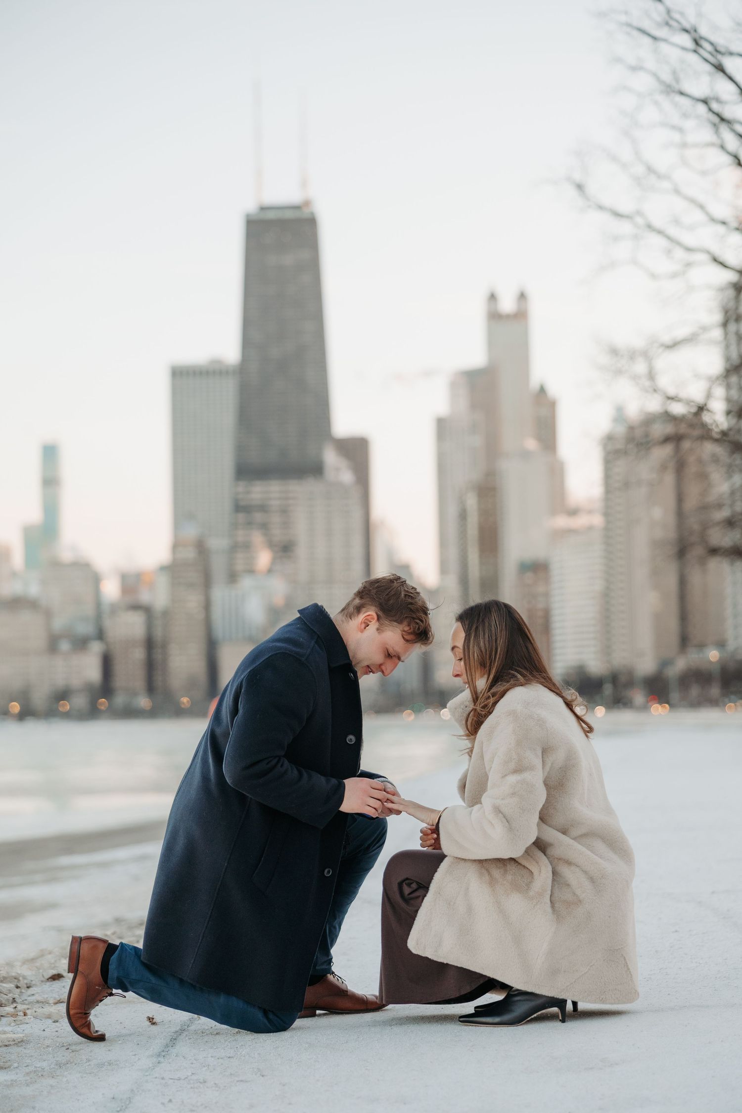Man proposes on one knee to woman in winter coats with Chicago skyline and John Hancock Center in background.