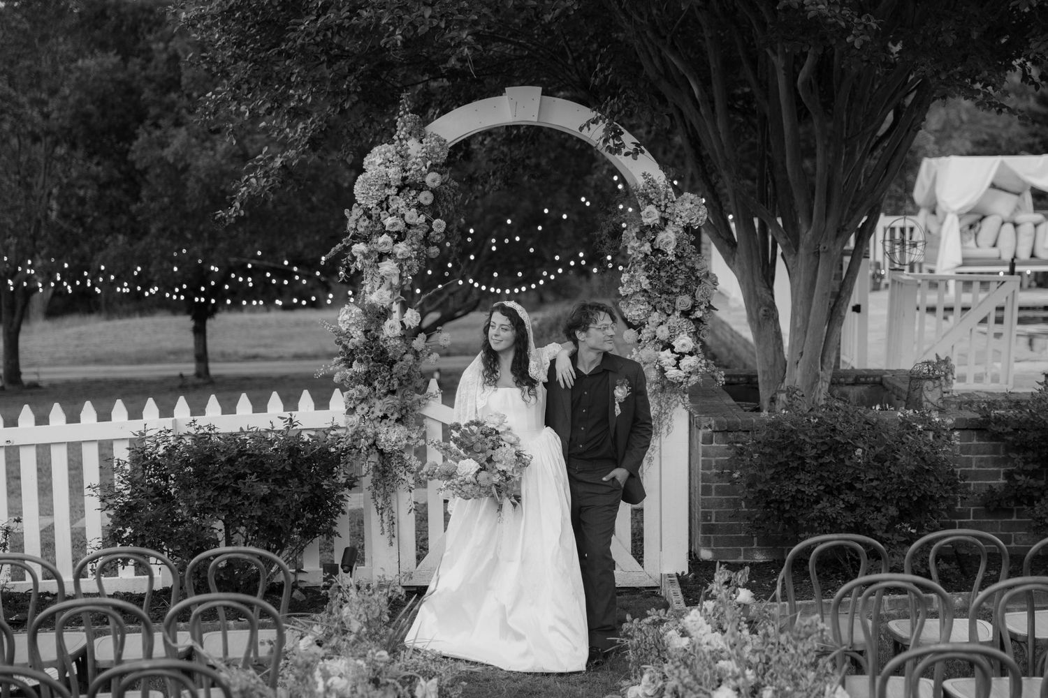 Black and white photo of bride and groom standing under floral arch at outdoor wedding ceremony.