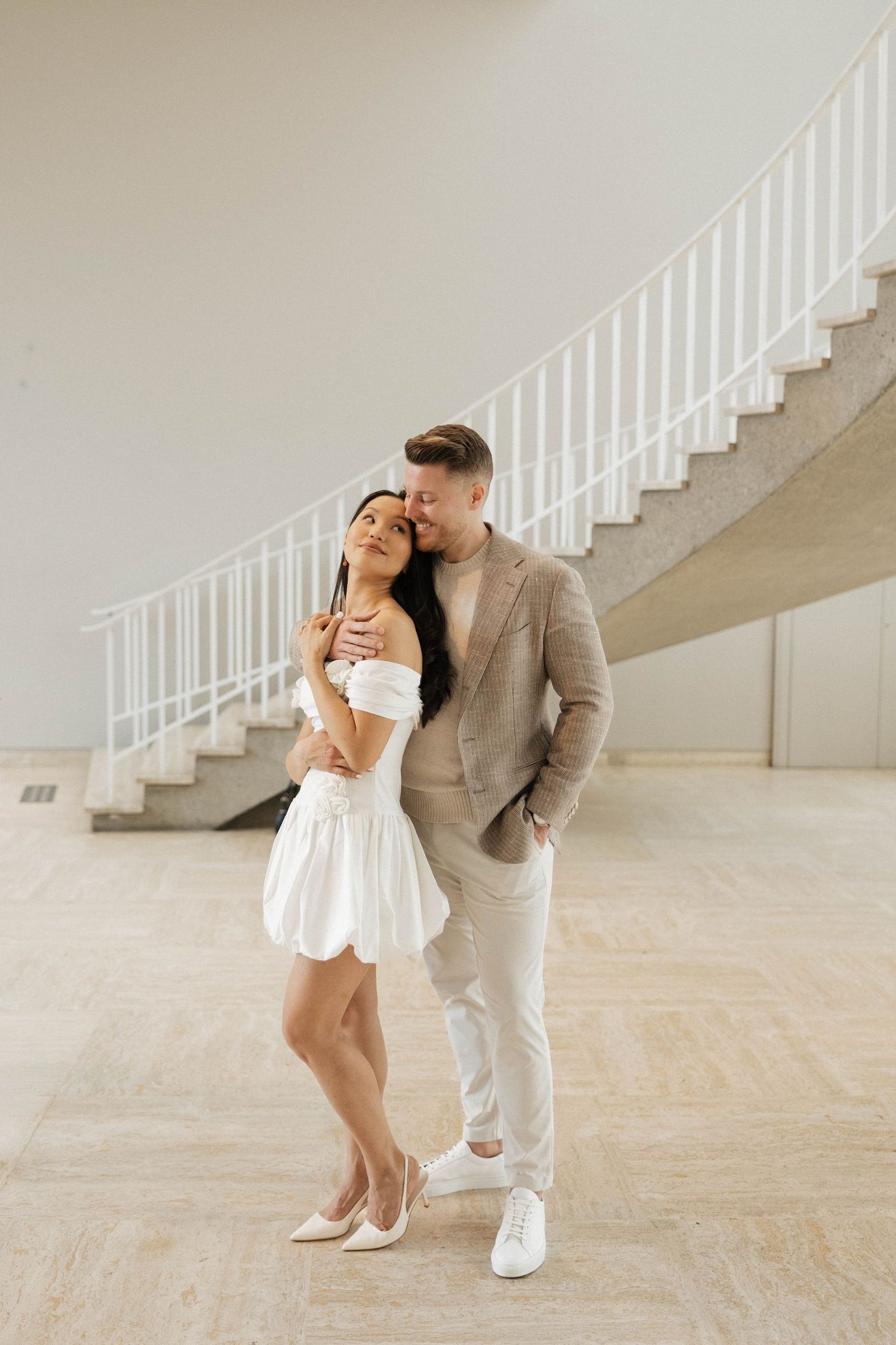 Couple embracing near elegant spiral staircase, woman in white mini dress and heels, man in beige suit.