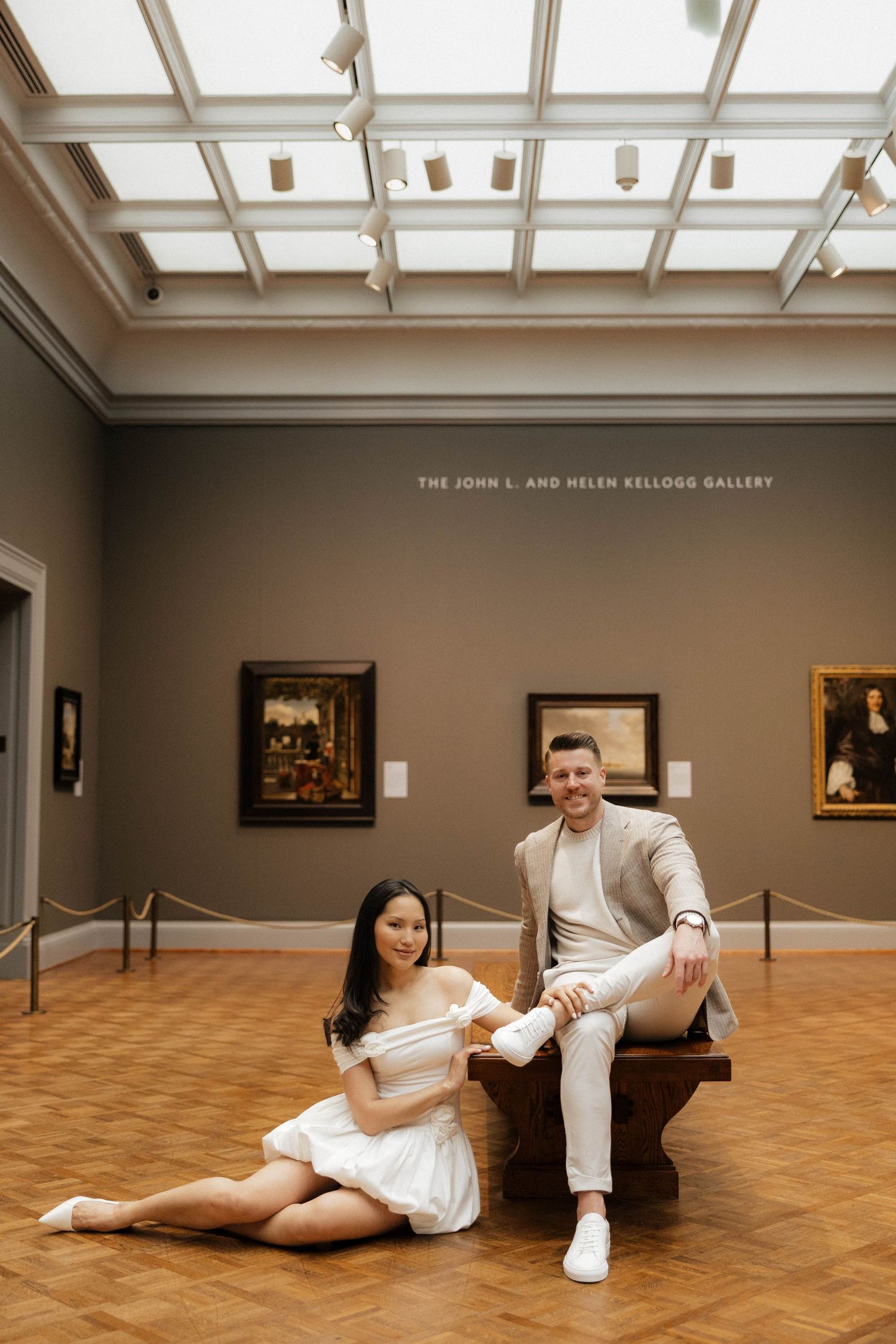 Couple posing in the John L. and Helen Kellogg Gallery, woman in white dress on floor, man in beige suit on bench.