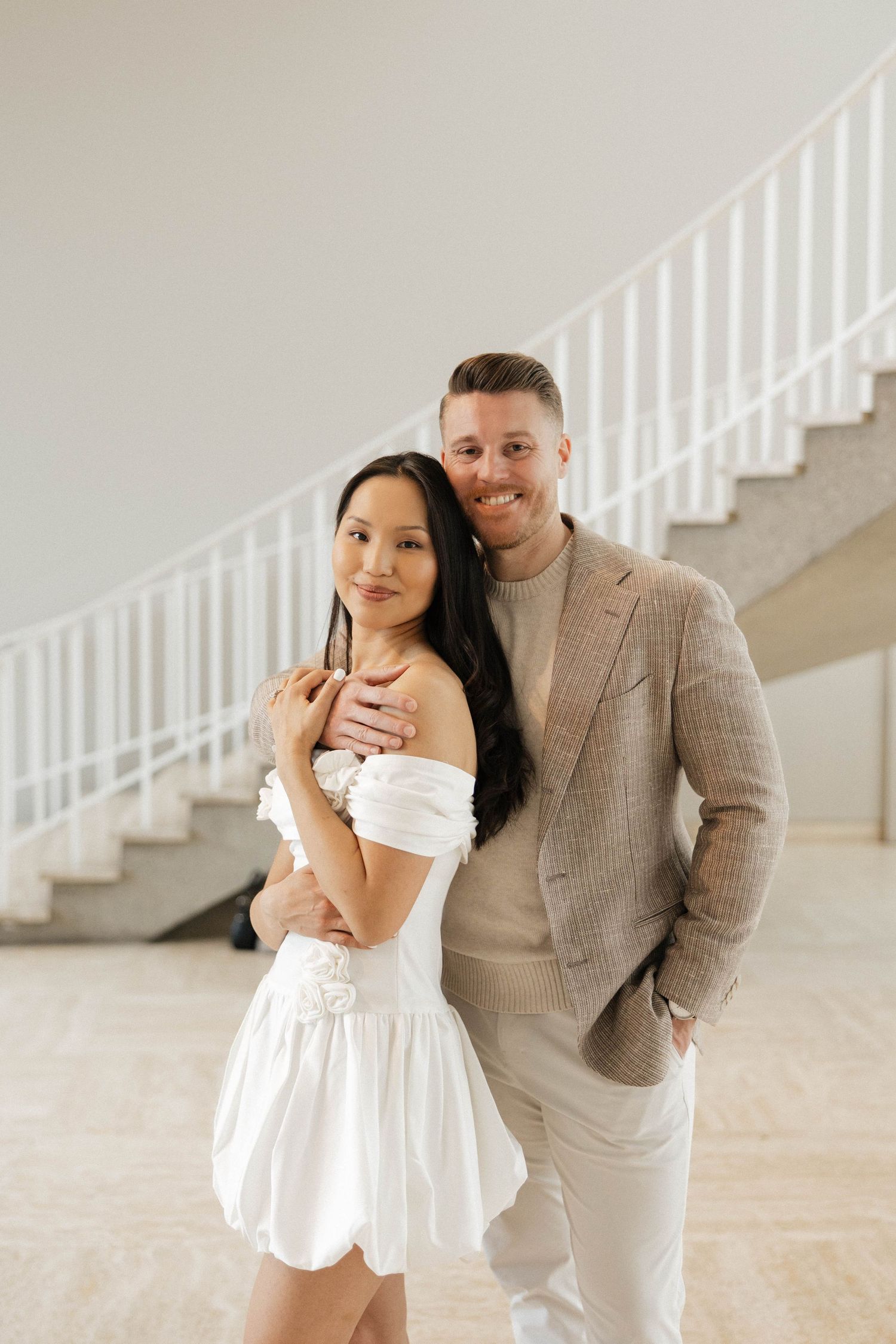 Couple posing elegantly, woman in white off-shoulder mini dress, man in beige blazer, embracing near white staircase.