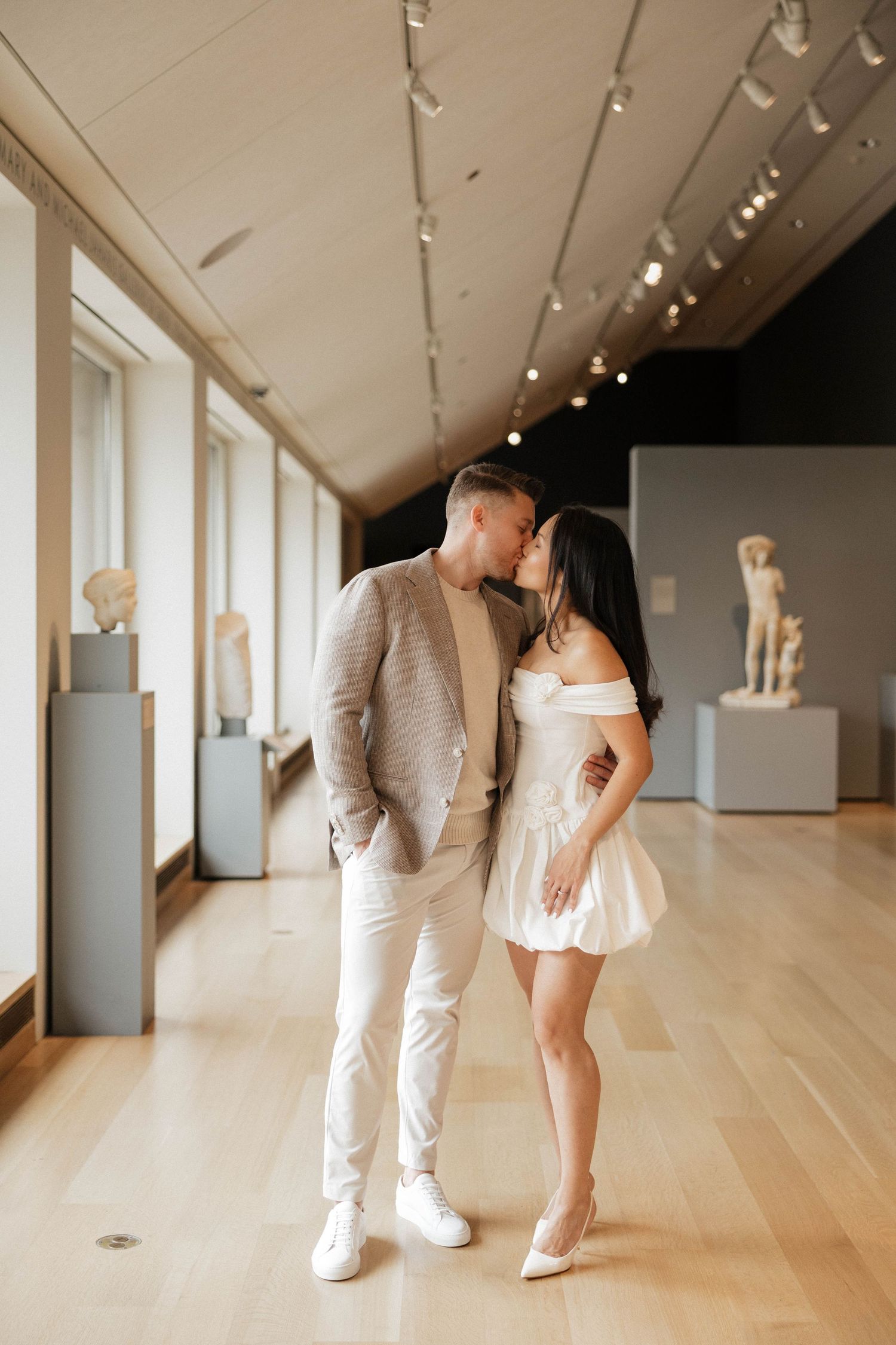 Couple sharing a romantic kiss in an art museum gallery with classical sculptures and warm lighting.