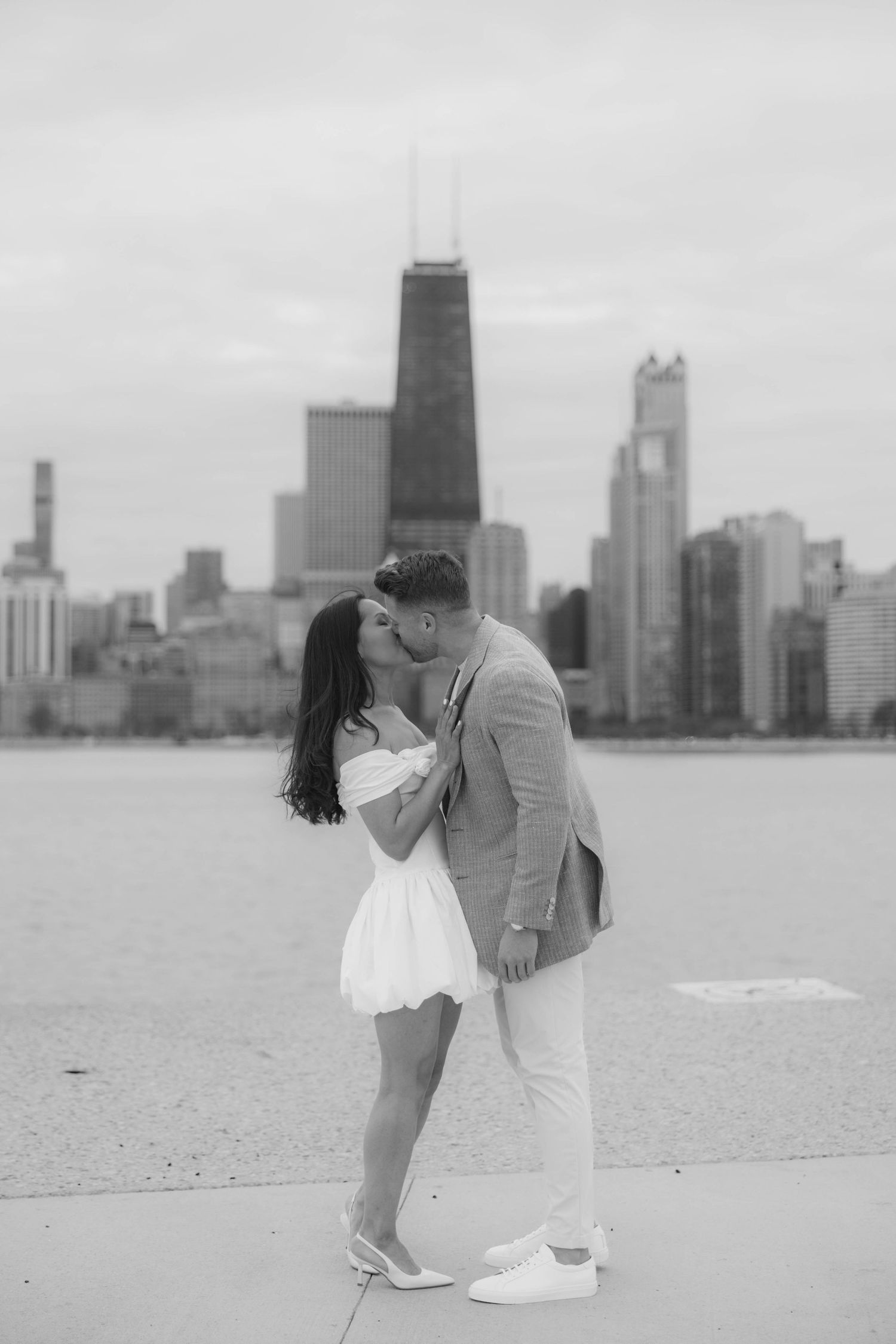 Black and white photo of couple kissing on Chicago lakefront with skyline in background.