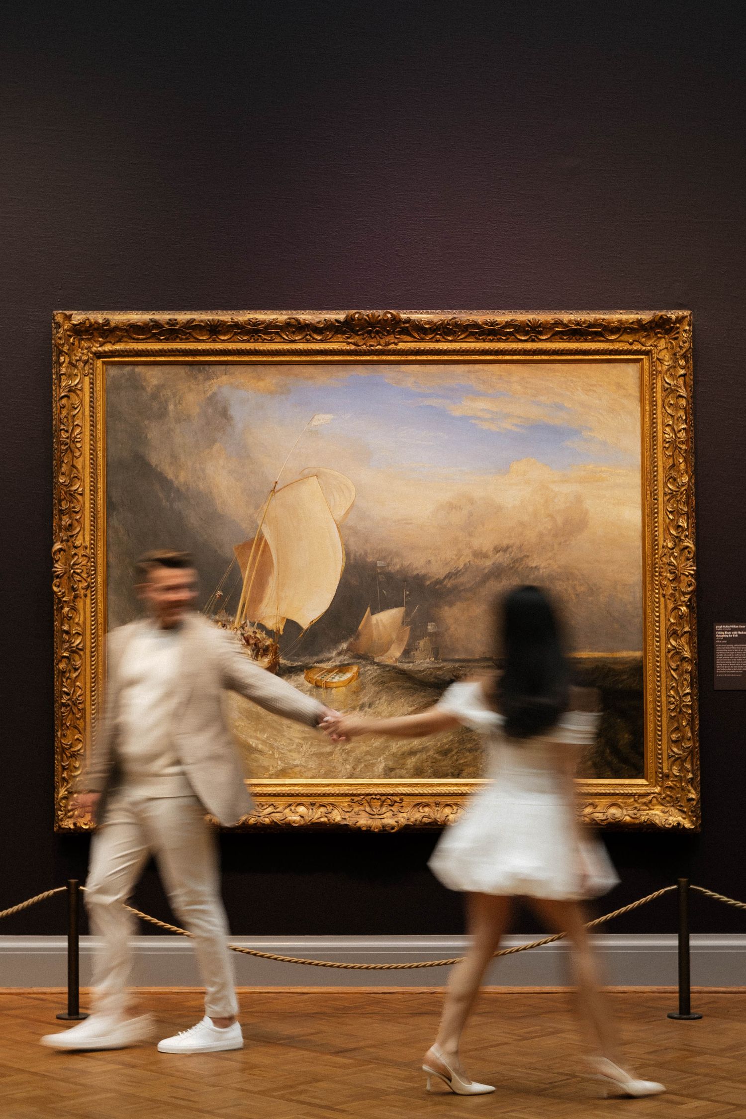 Couple holding hands walking past a large J.M.W. Turner seascape painting in a golden frame at an art museum.