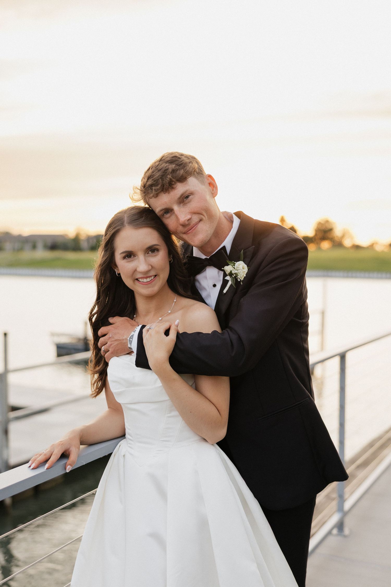 Bride and groom embracing on a waterfront dock at sunset, groom in black tuxedo, bride in white gown.
