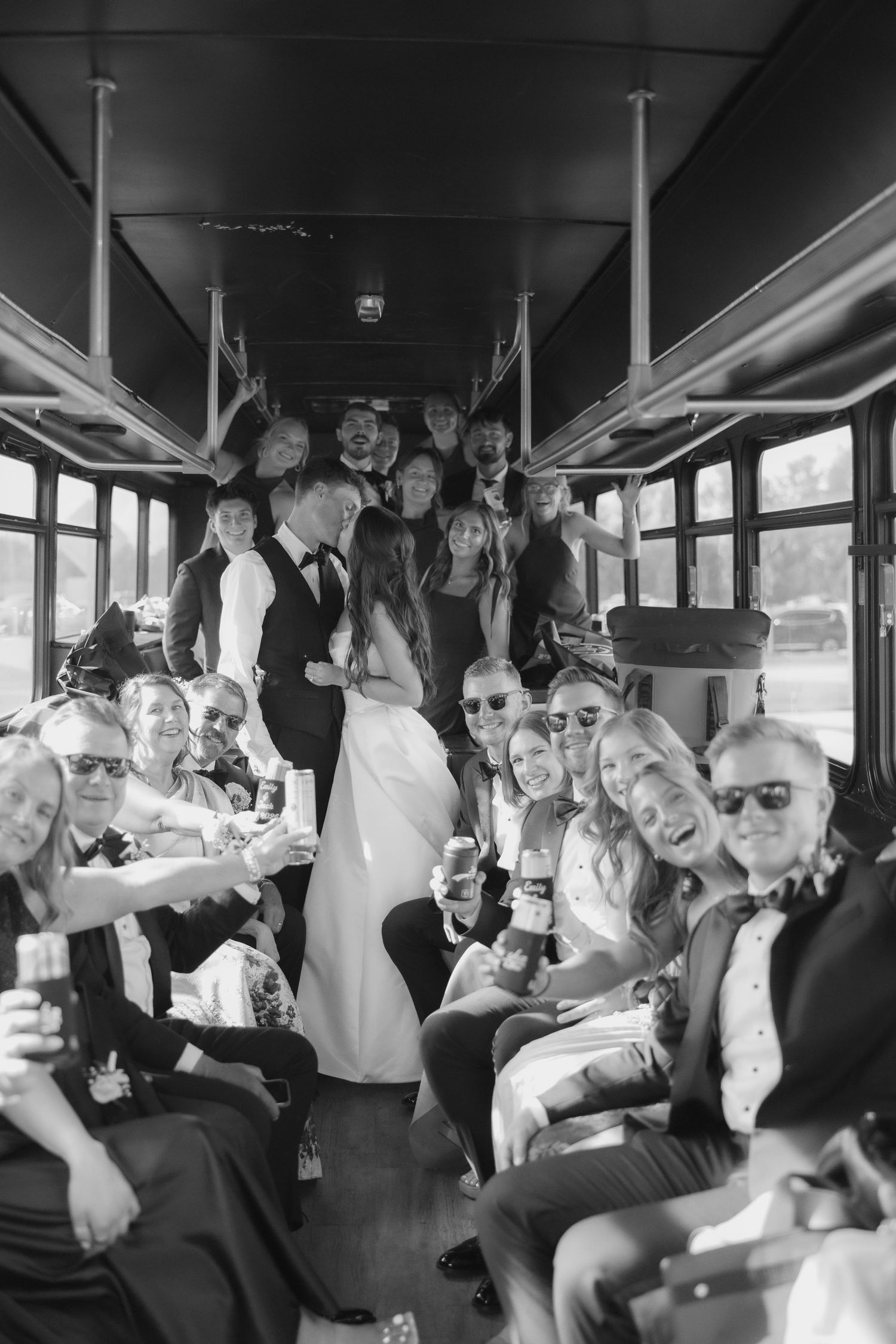 Black and white photo of bride and groom kissing surrounded by cheering wedding guests on a party bus.