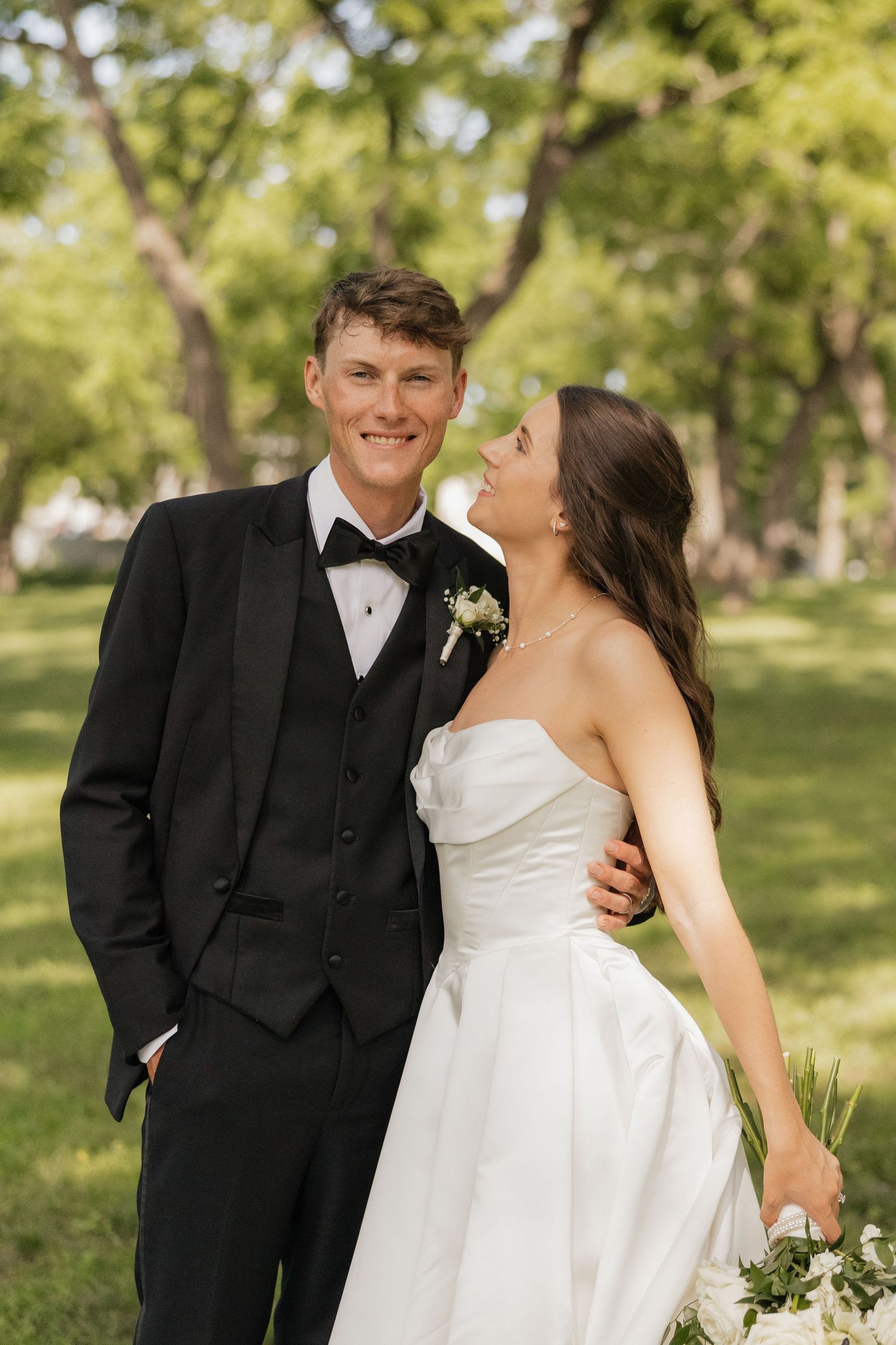 A smiling groom in a black tuxedo stands with his bride in a white strapless gown in a lush green park.