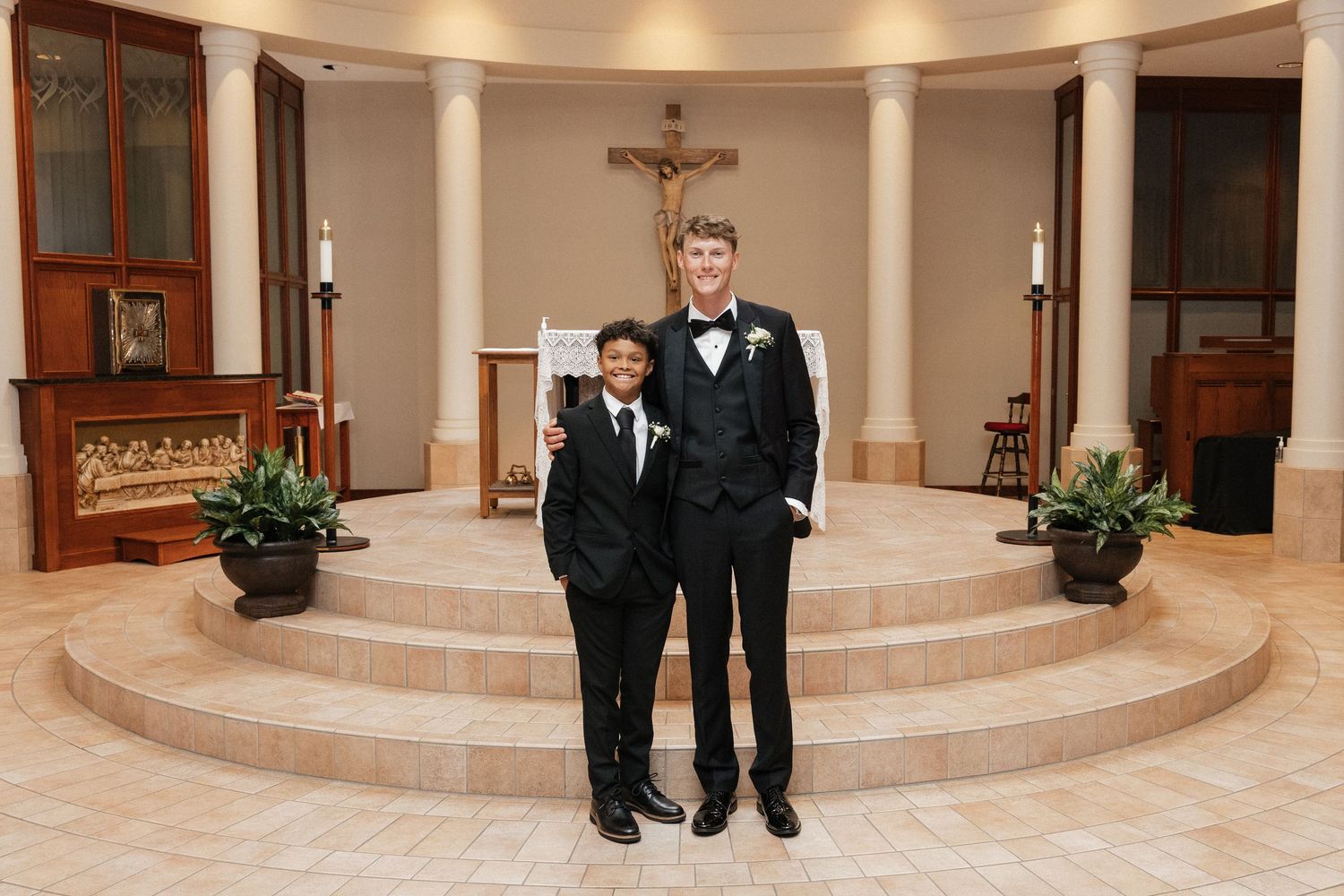 Two boys in formal black suits posing together inside a Catholic church with a crucifix altar backdrop.