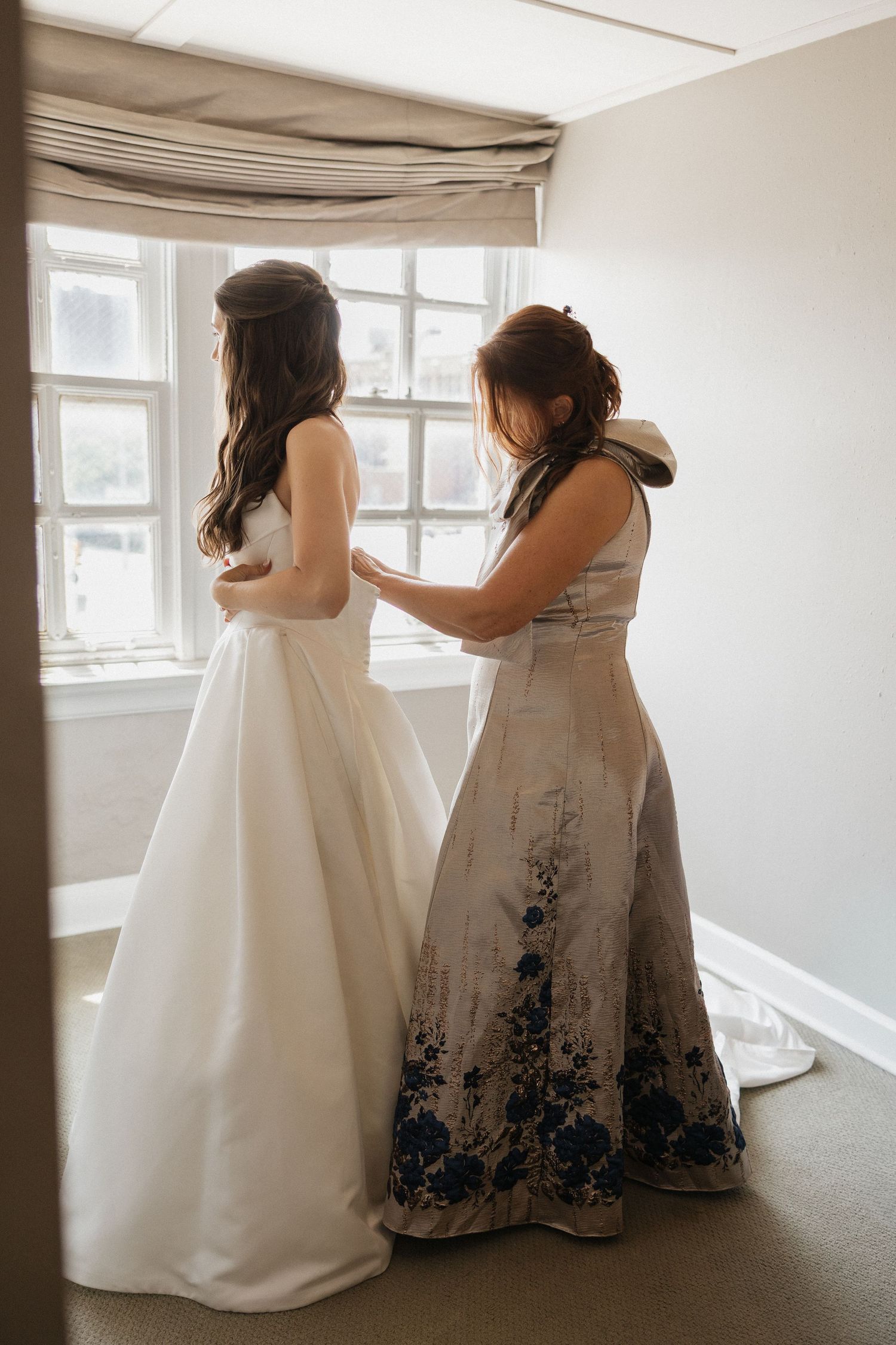 A bride in a white gown stands by a window as a woman in a floral dress helps fasten her wedding dress.