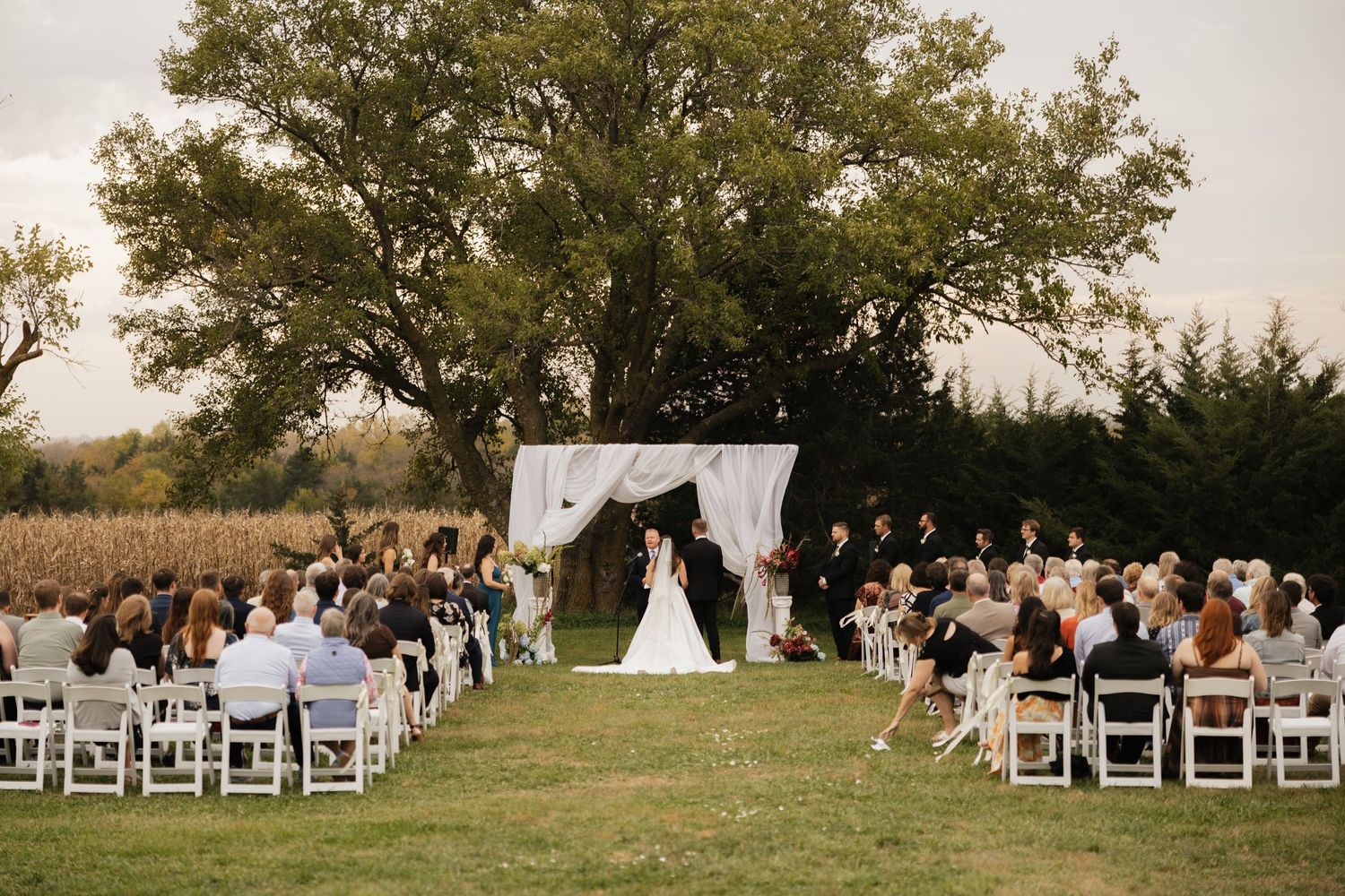 Bride and groom exchange vows under a draped white canopy as seated guests watch at an outdoor wedding ceremony at sunset.