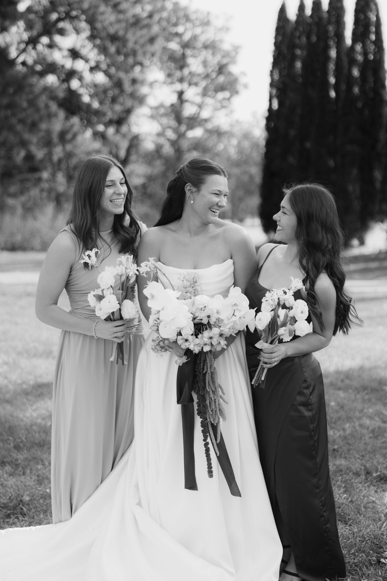 Black and white photo of a smiling bride in a strapless gown laughing with two bridesmaids holding white floral bouquets outdoors.