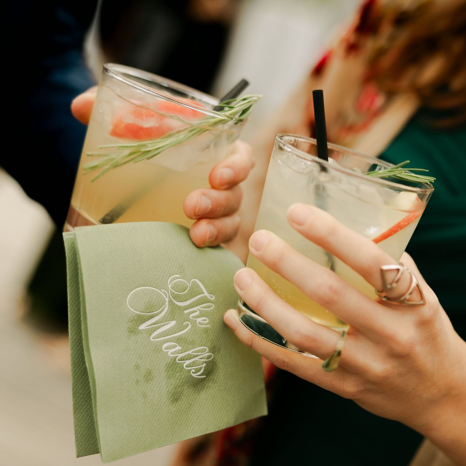 Two people toast with citrus cocktails garnished with rosemary, holding a green personalized cocktail napkin at a wedding reception.