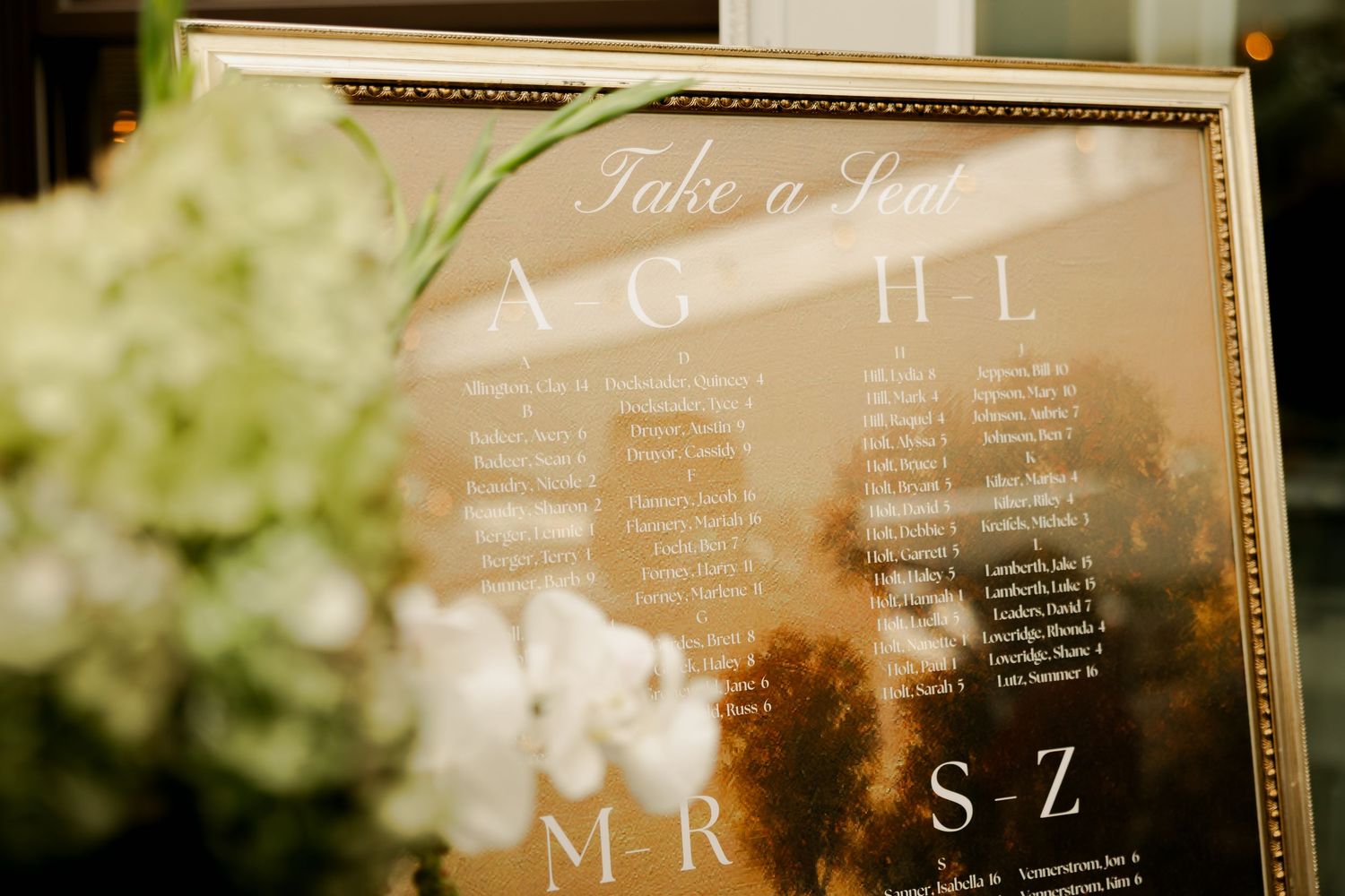 Gold-framed mirror wedding seating chart reading Take a Seat, with white hydrangea and orchid flowers beside it.