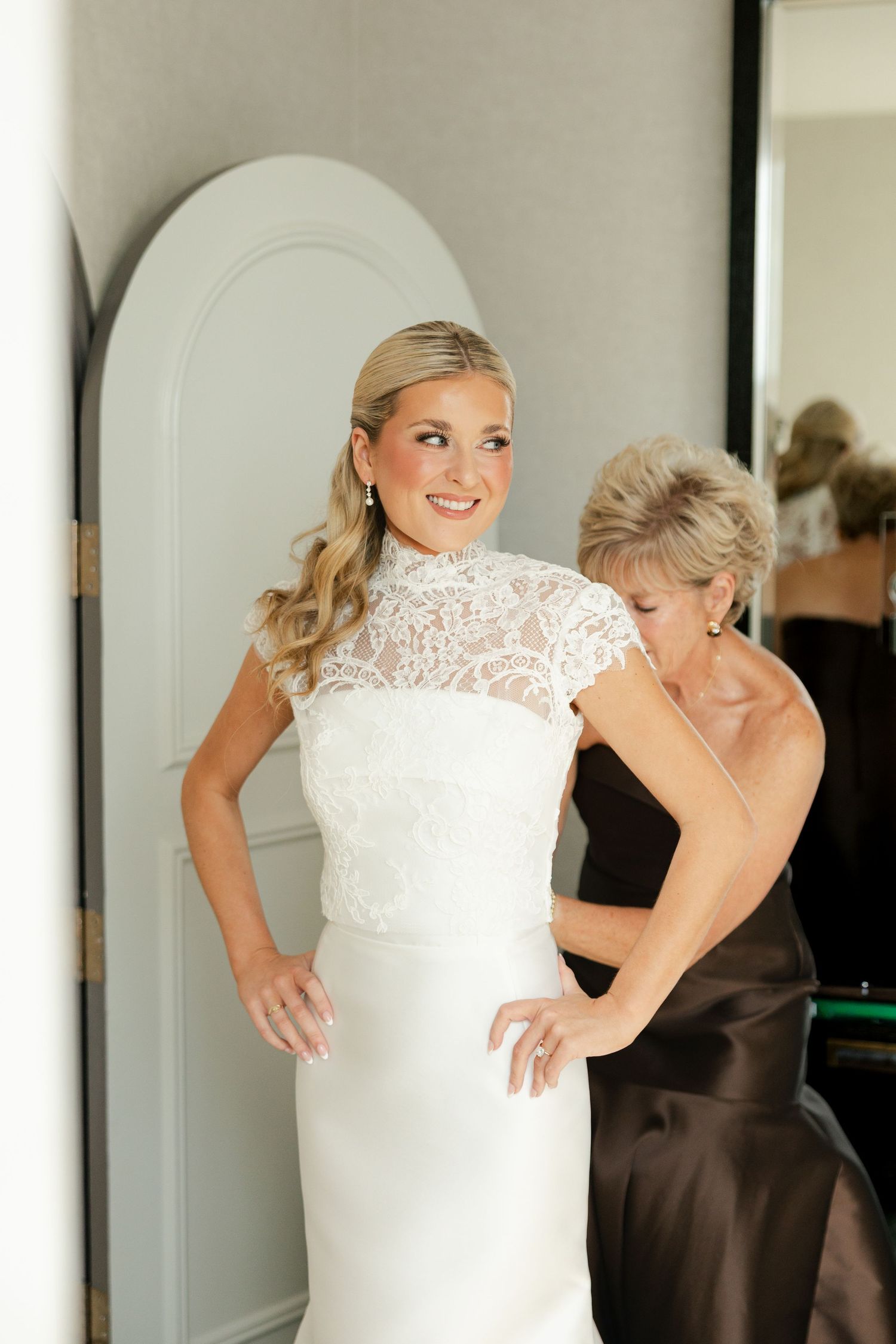 Smiling bride in an elegant white lace high-neck wedding gown gets help with her dress from a woman in a brown satin dress.