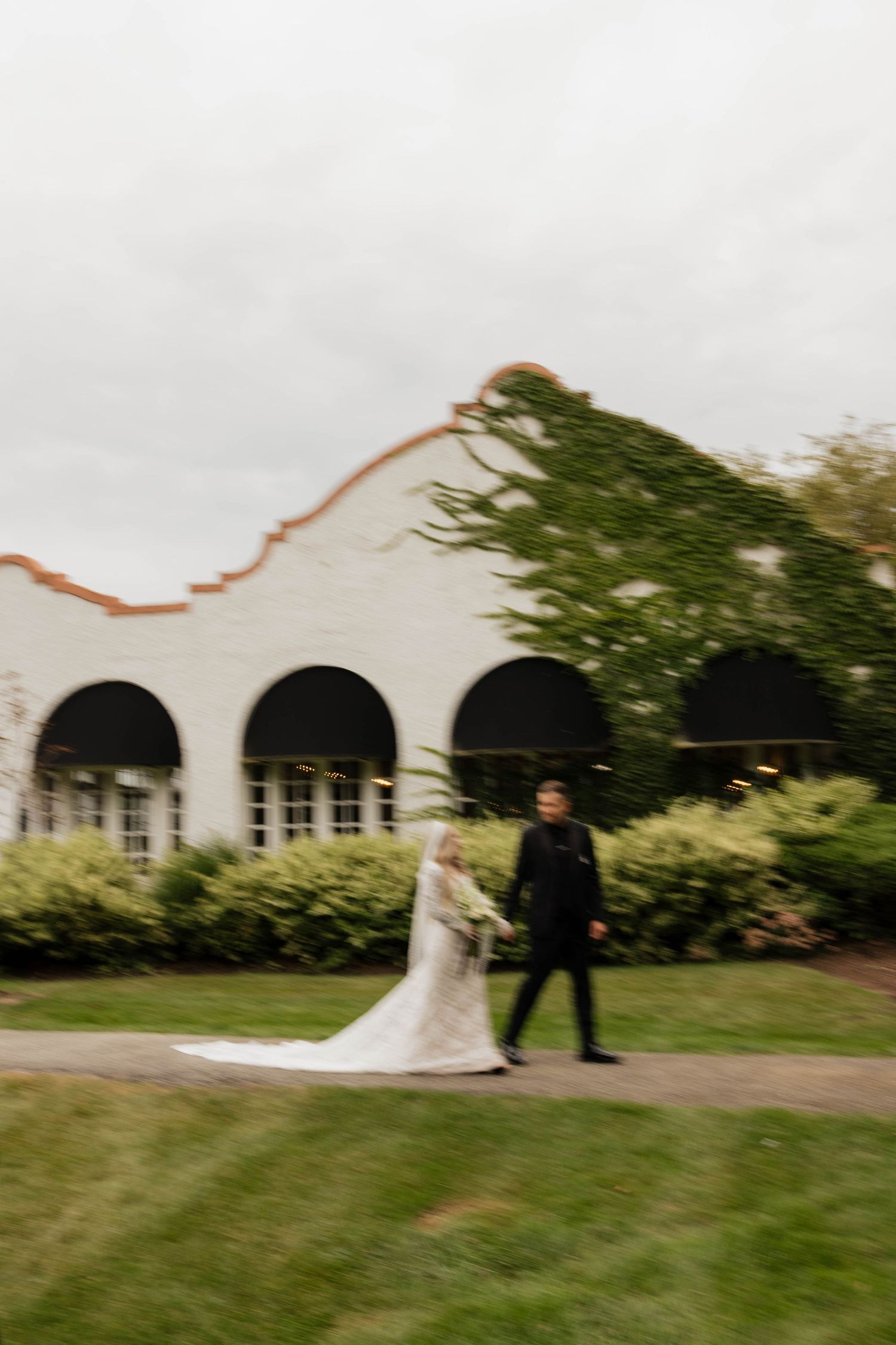 Bride in a lace gown and veil walks hand in hand with a groom in black past an ivy-covered white building with arched windows.