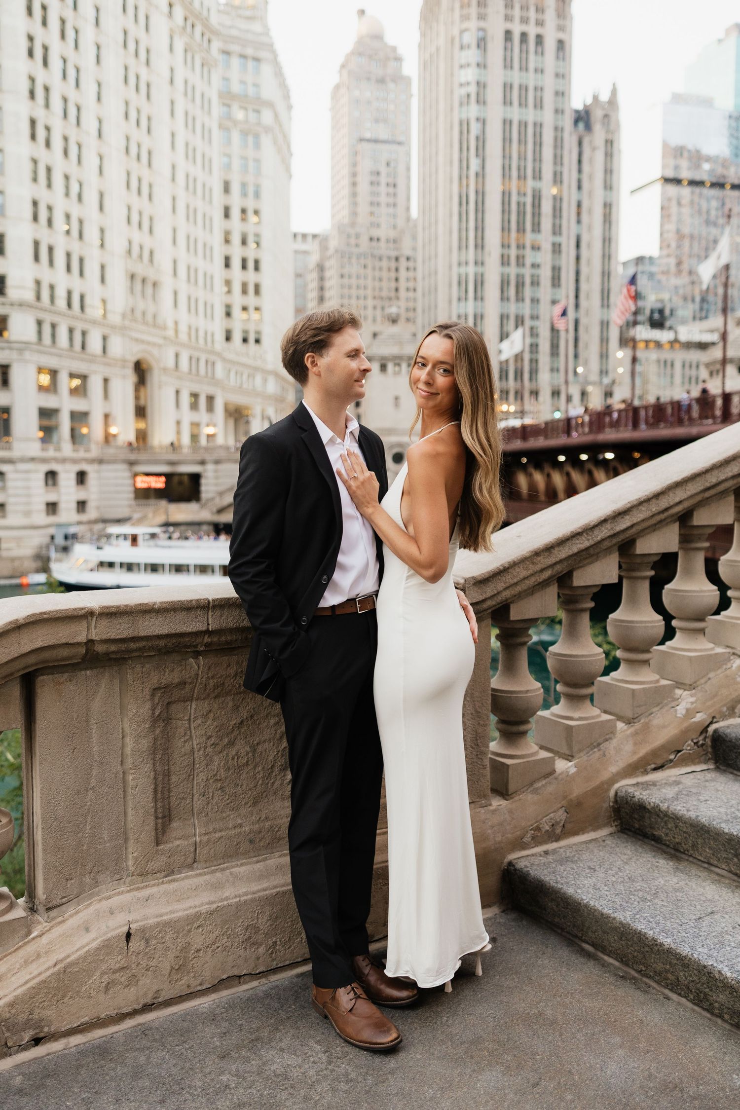 Couple embraces on Chicago Riverwalk bridge, woman in white gown leans against man in black suit, skyscrapers behind.