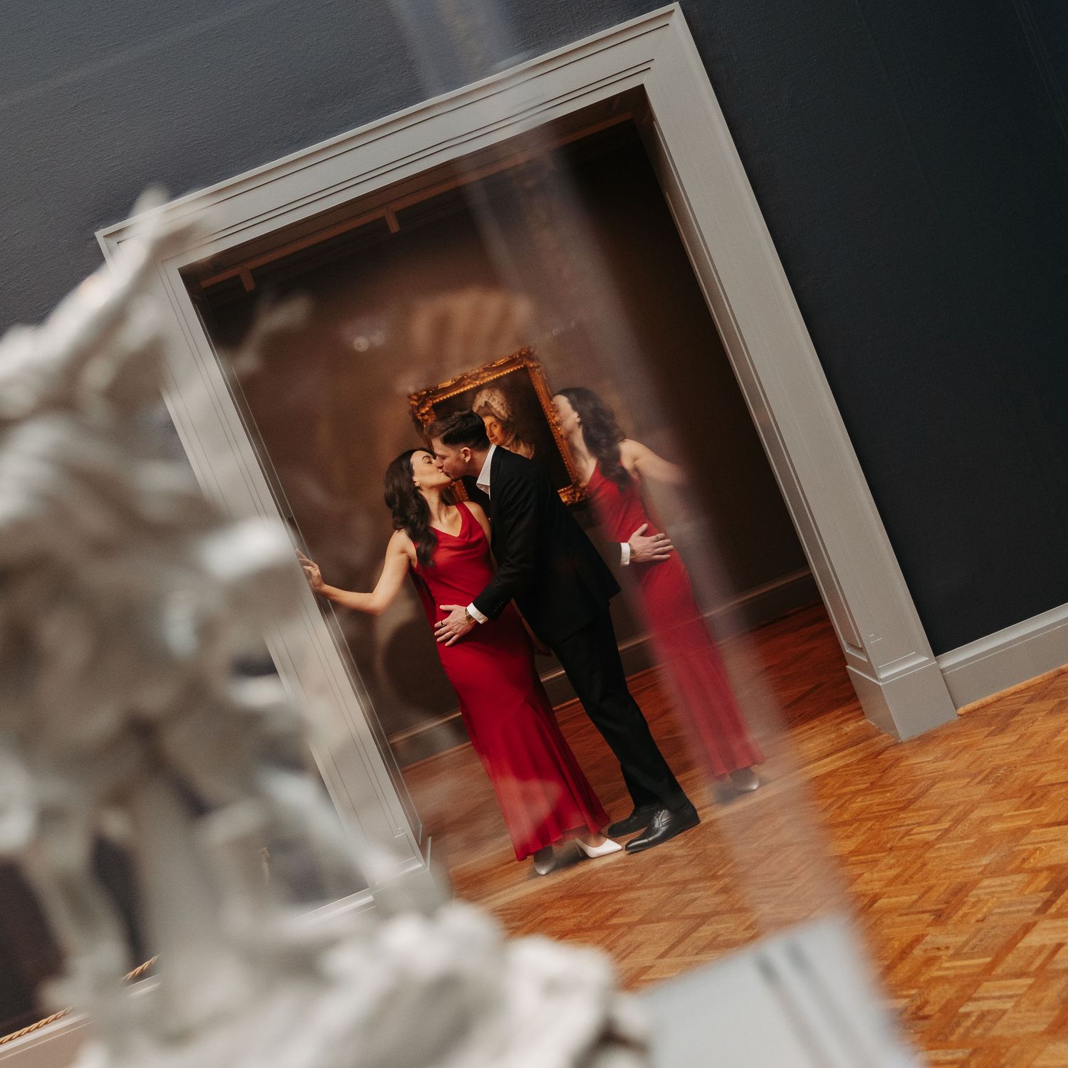 Couple in formal wear embracing in a museum doorway, reflected in glass, with a white sculpture in the foreground.