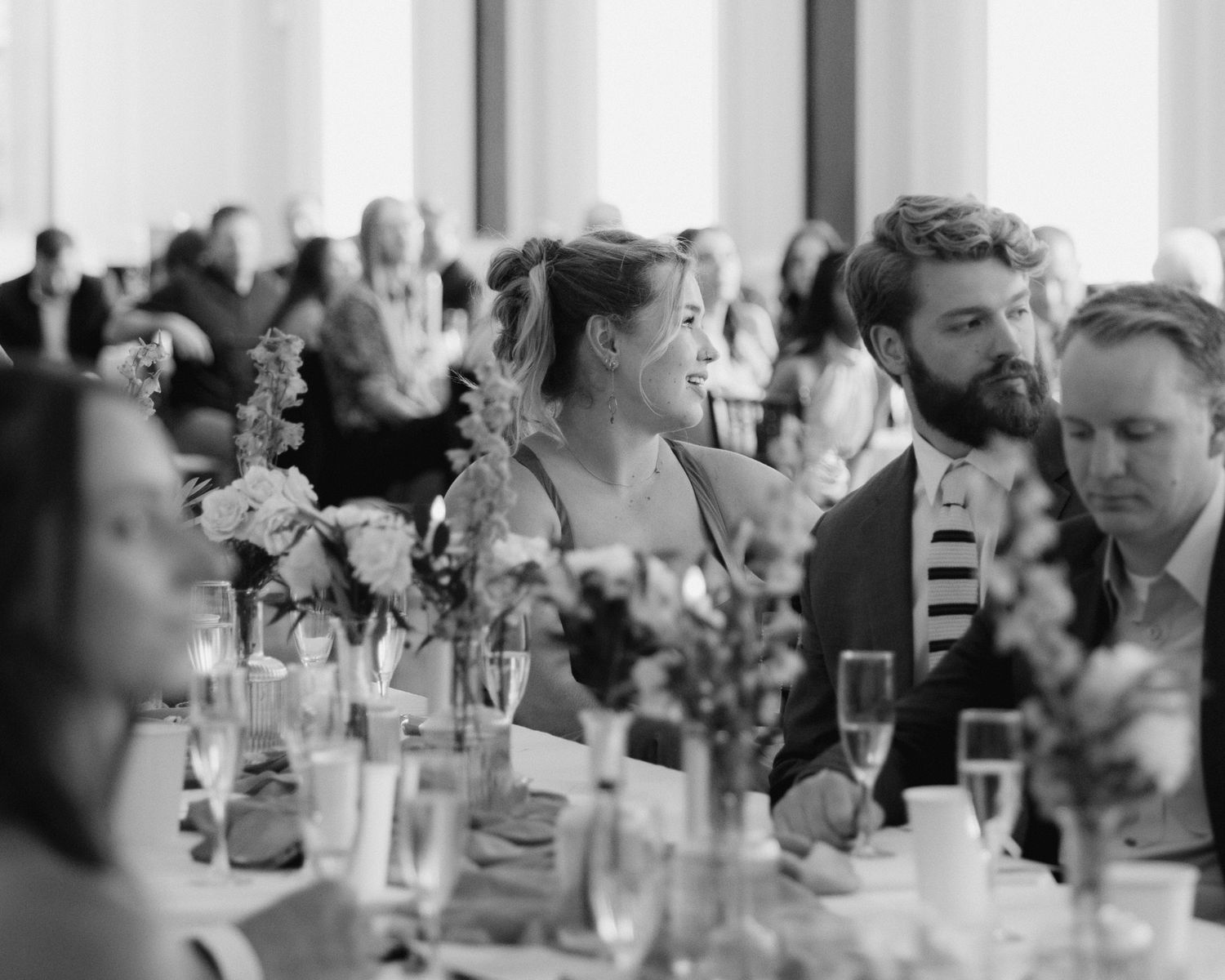 Wedding guests sit at long tables during an elegant reception dinner with soft lighting and floral centerpieces.