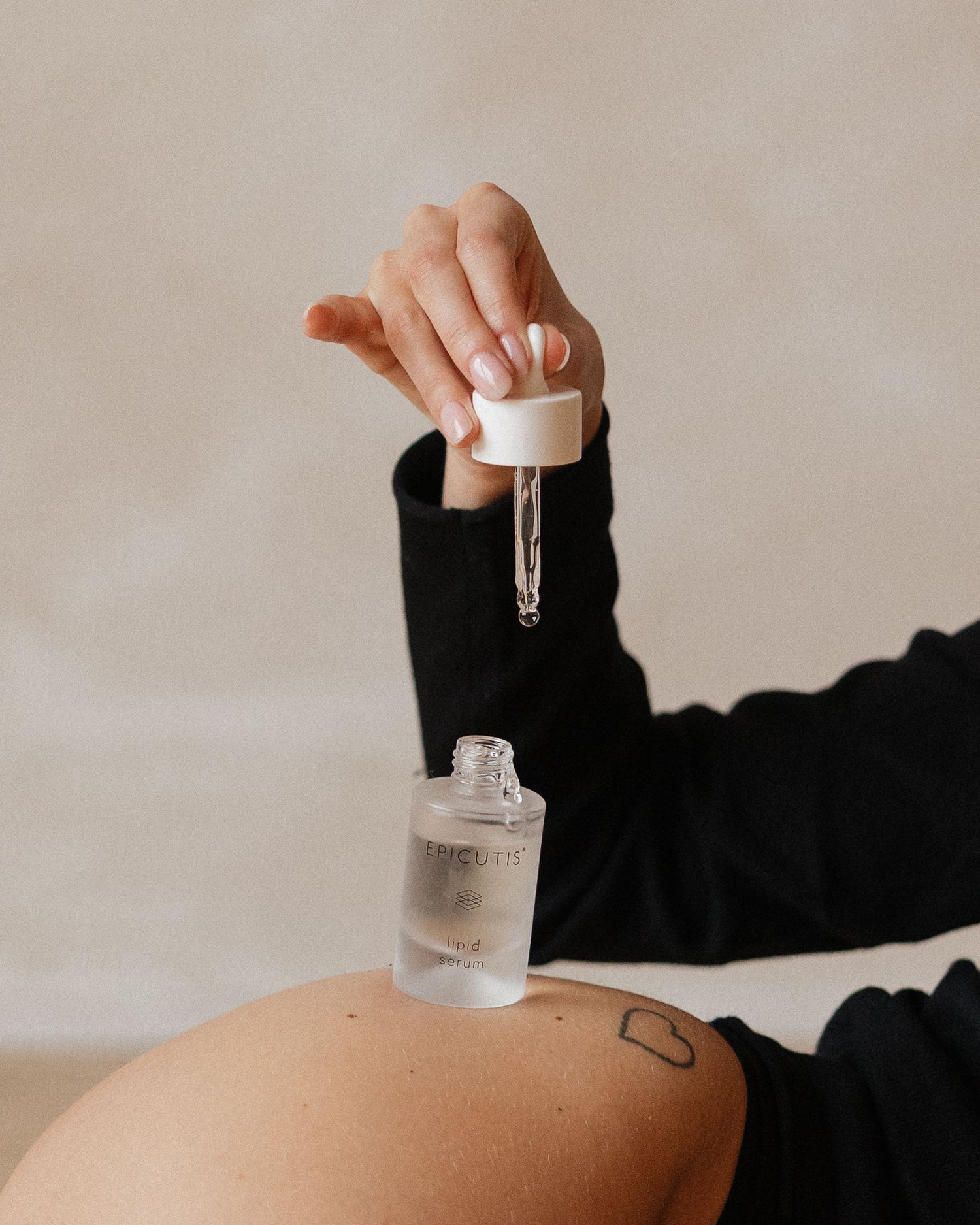 A hand holding a dropper of skincare serum over bare skin with a small tattoo visible.