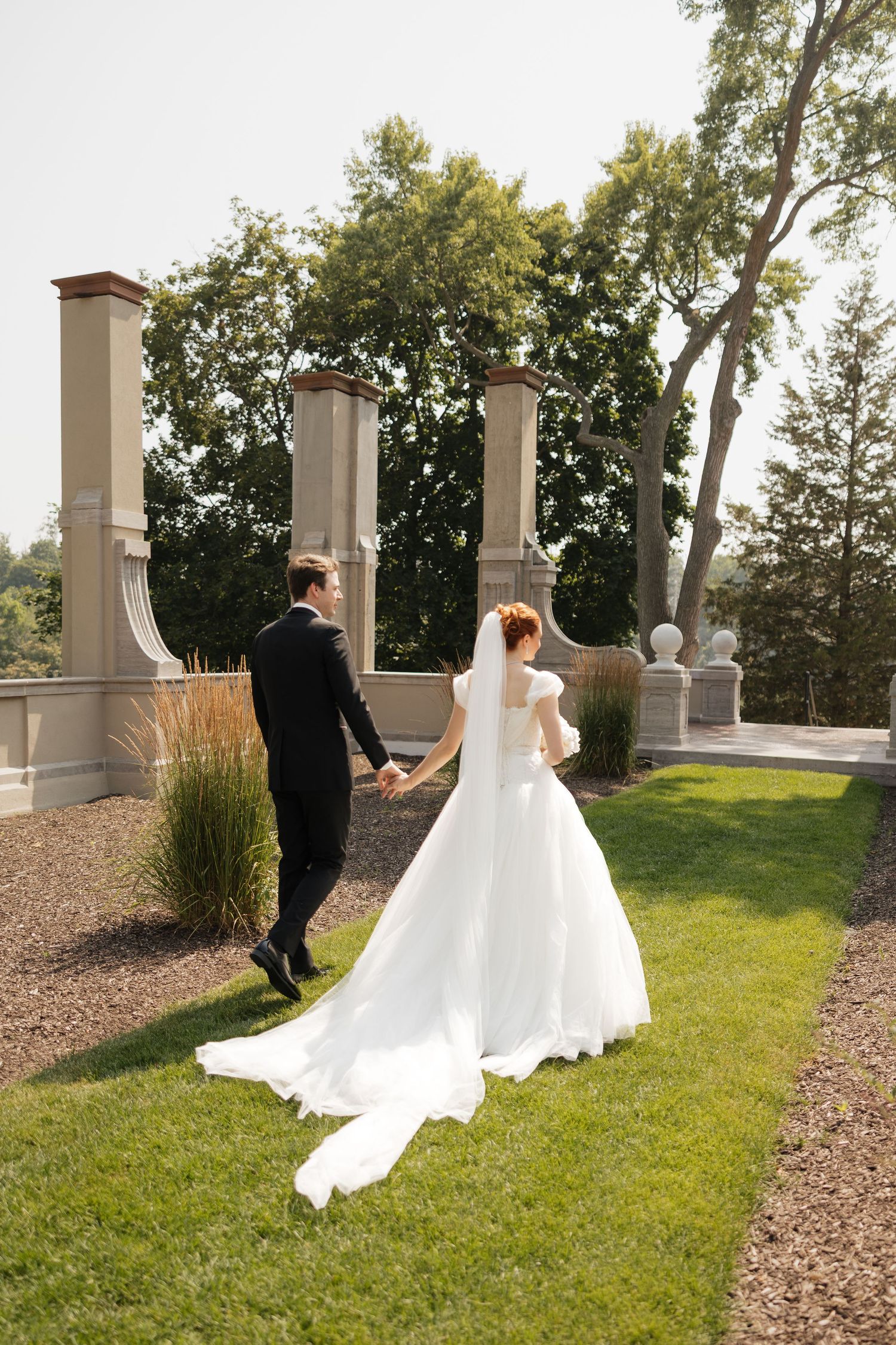 Bride and groom walk hand in hand through elegant garden venue, bride wearing flowing white gown with long veil and train.