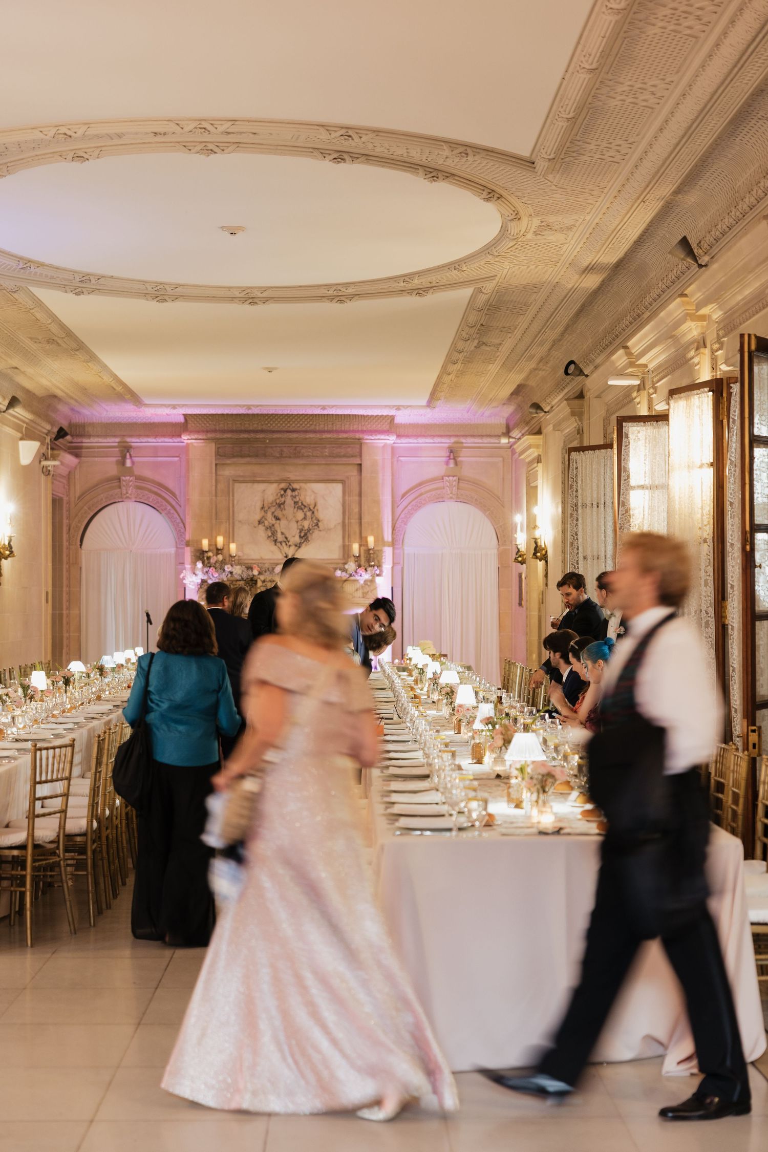 Elegant wedding reception in ornate ballroom with long dining table, gold chairs, and guests in formal attire.