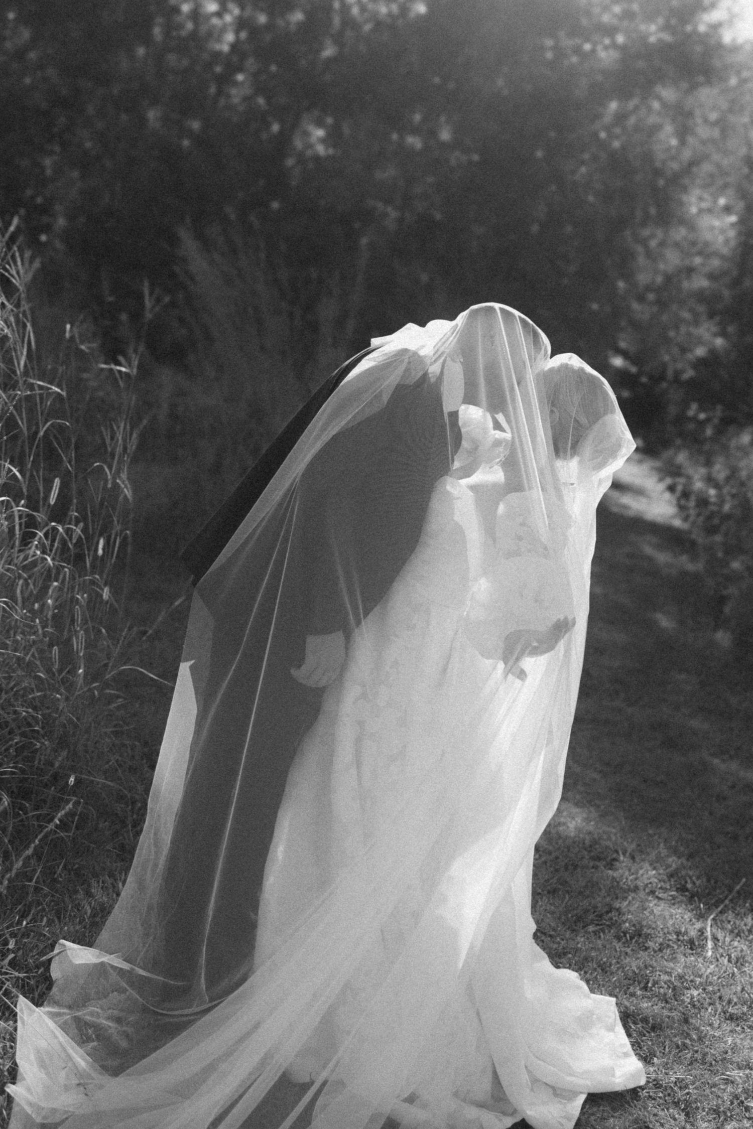 A flowing white wedding veil billows in the wind against a dark forest background in black and white photography.