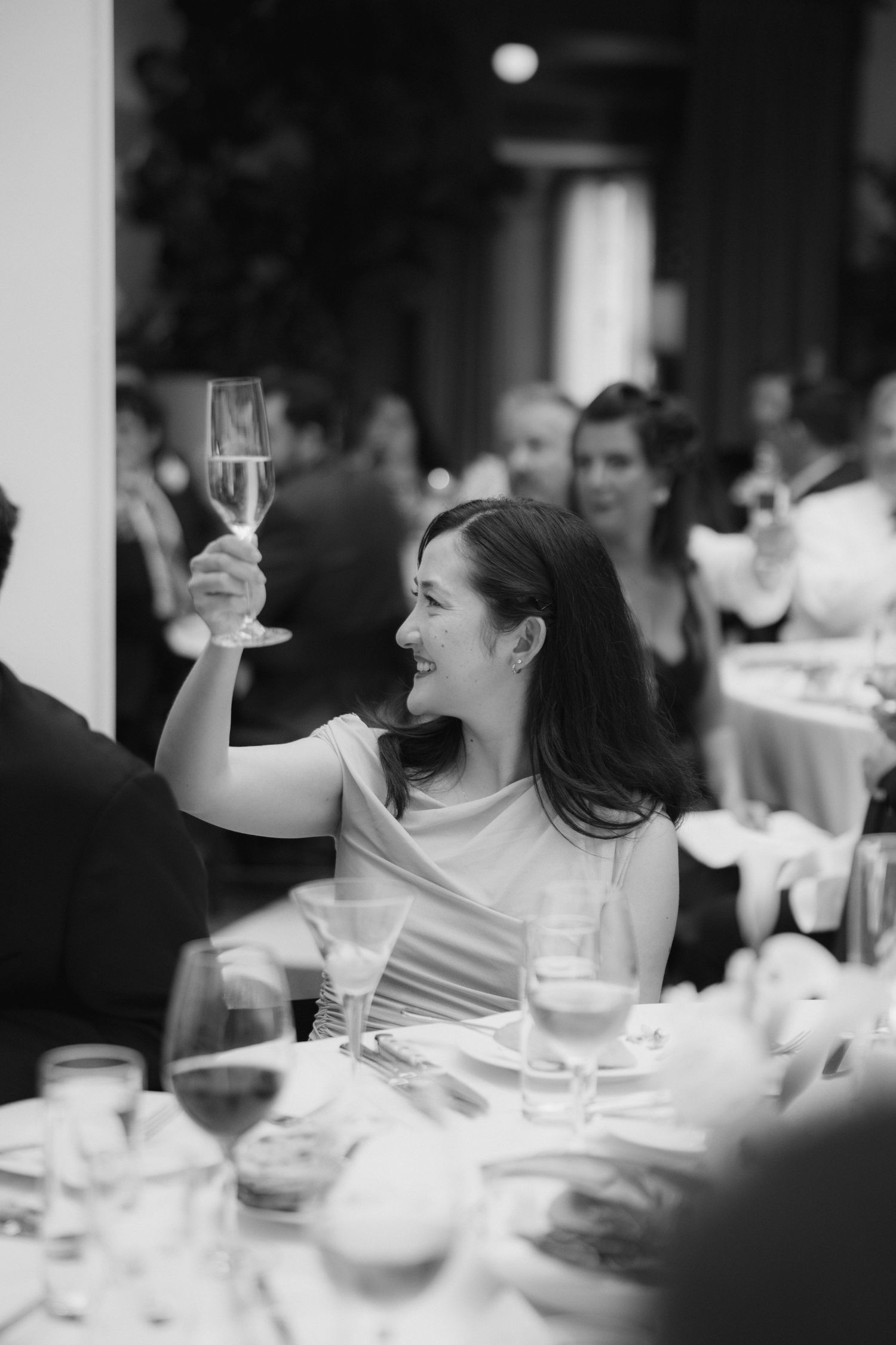 Black and white photo of smiling wedding guest raising champagne glass in a toast at reception.