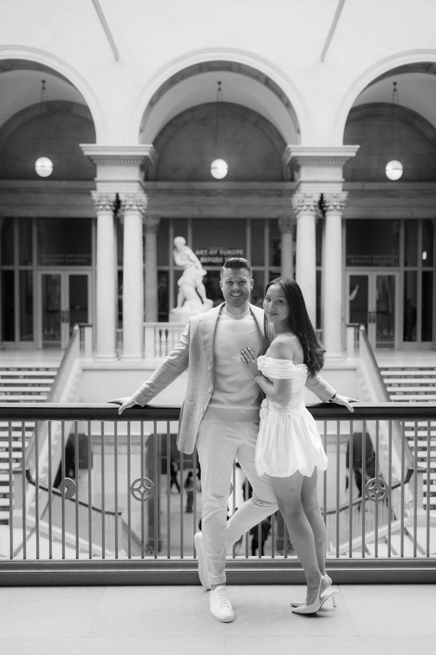 Black and white photo of elegant couple posing on balcony inside grand classical building with arches and columns.