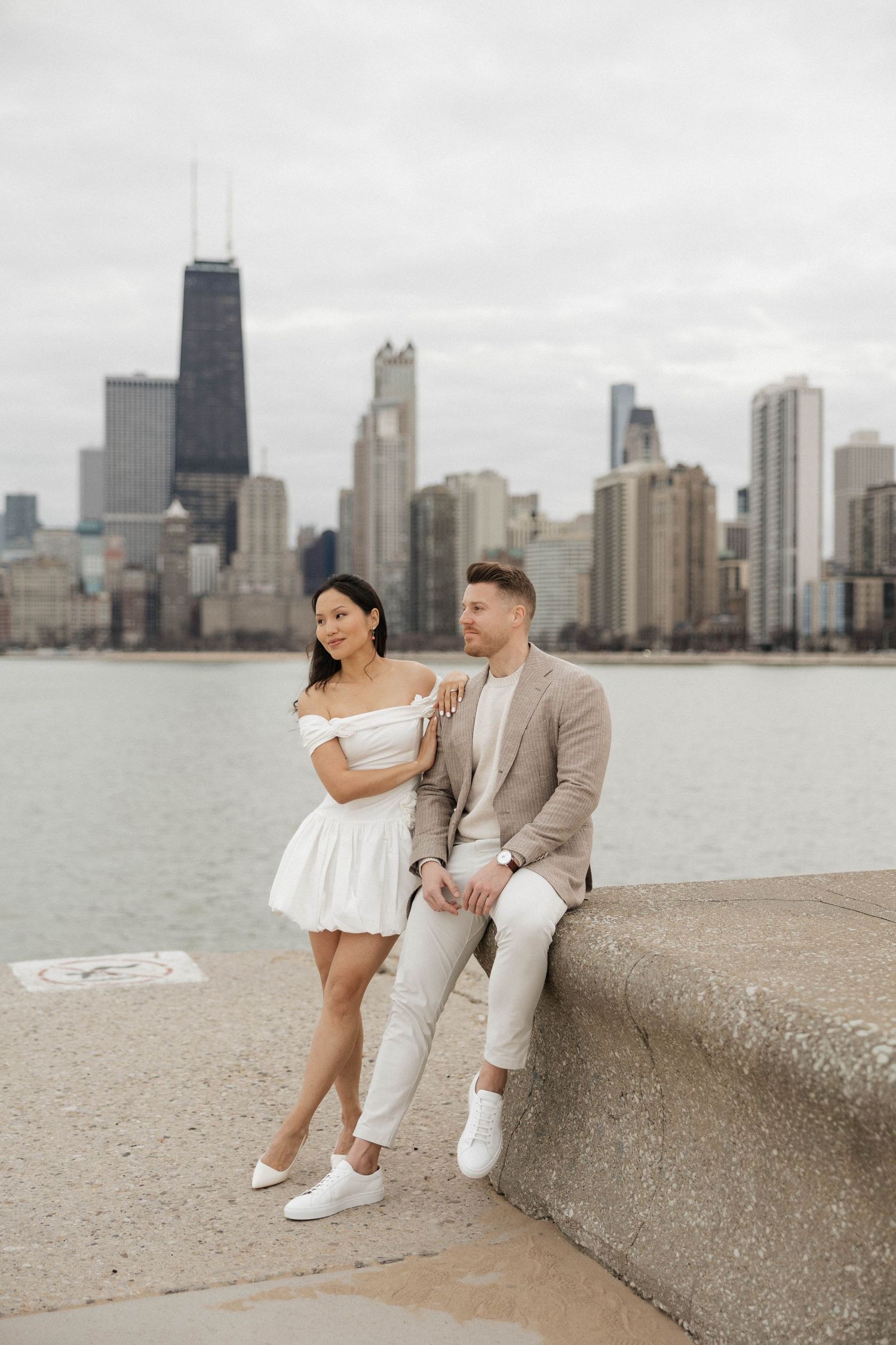 Couple posing by Lake Michigan with Chicago skyline backdrop, woman in white dress, man in beige suit.